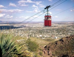 A red tram car hangs from a dark wire, El Paso is visible below in a wide valley expanse