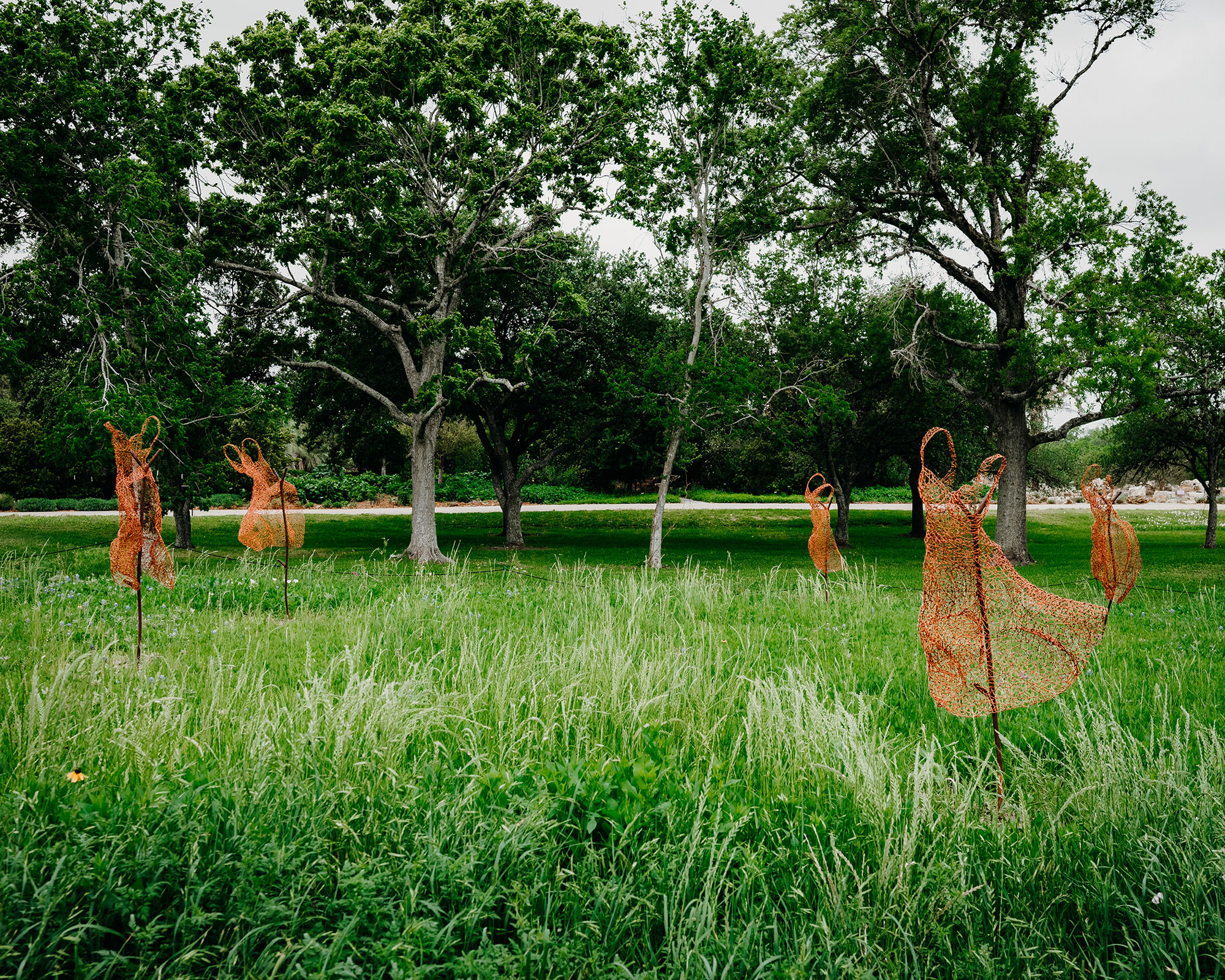 Five sculptures, resembling dancing human forms made of red wires, are placed several feet from each other in a grassy area with tall leafy green trees in background.