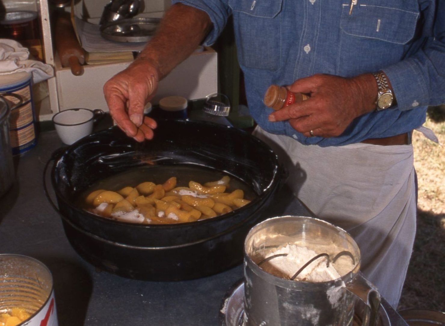 A man in a cowboy hat lays slices of canned peaches in a Dutch oven. He's in a cluttered room with storage boxes lining the wall behind him.