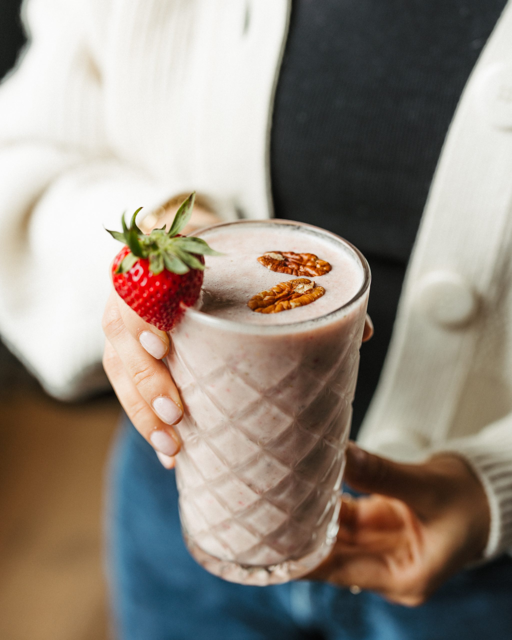 A person holds a glass of strawberry milkshake with two pecans floating on top and a strawberry on the rim