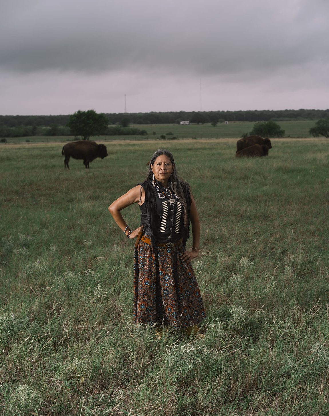 A woman with long gray and brunette hair dressed in a clothing with Native American patterns and jewlery stands stoically in a field with several buffalo in the background.