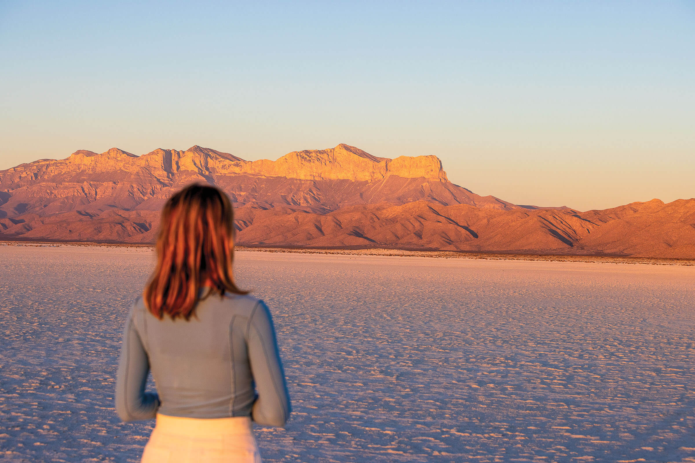 A woman in a grey long-sleeved shirt faces away from the camera and overlooks a flat, sandy dune with a mountain in the background. The sun, which has either just set or risen, turns the mountain dusky yellow and purple.