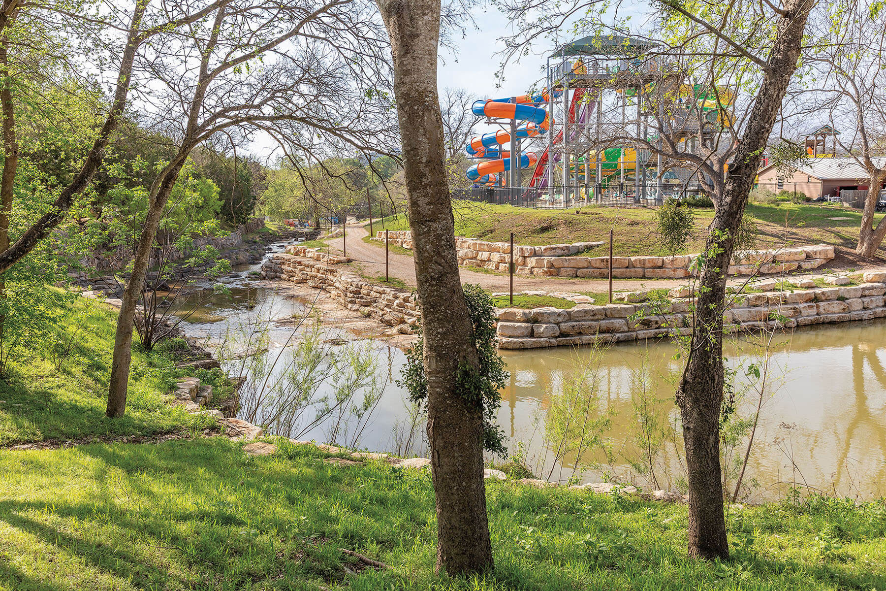 Bright orange and blue plastic waterslides are visible in the background of a river area with green grass and trees