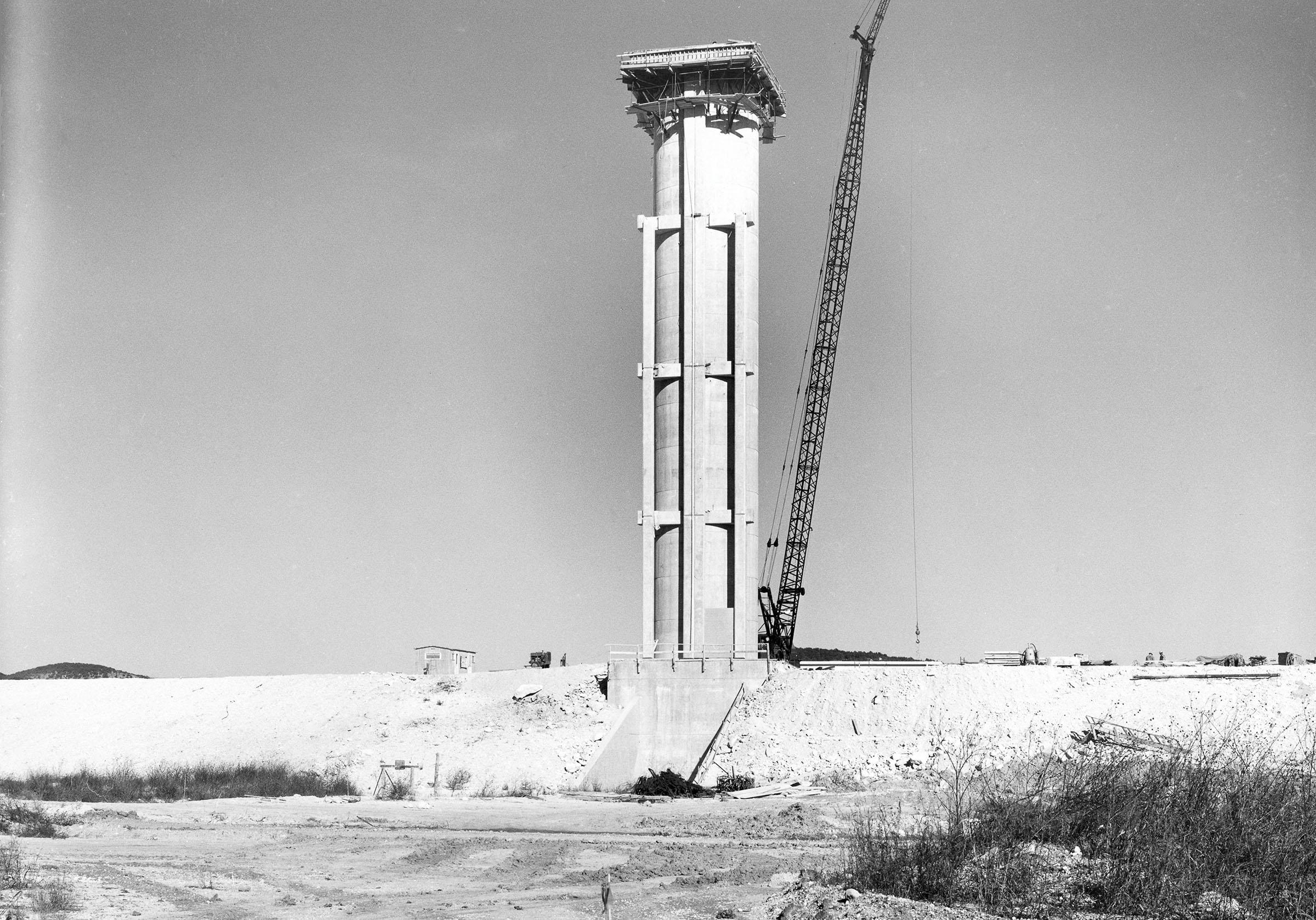 A black and white image of large construction vehicles and a tower on the shore