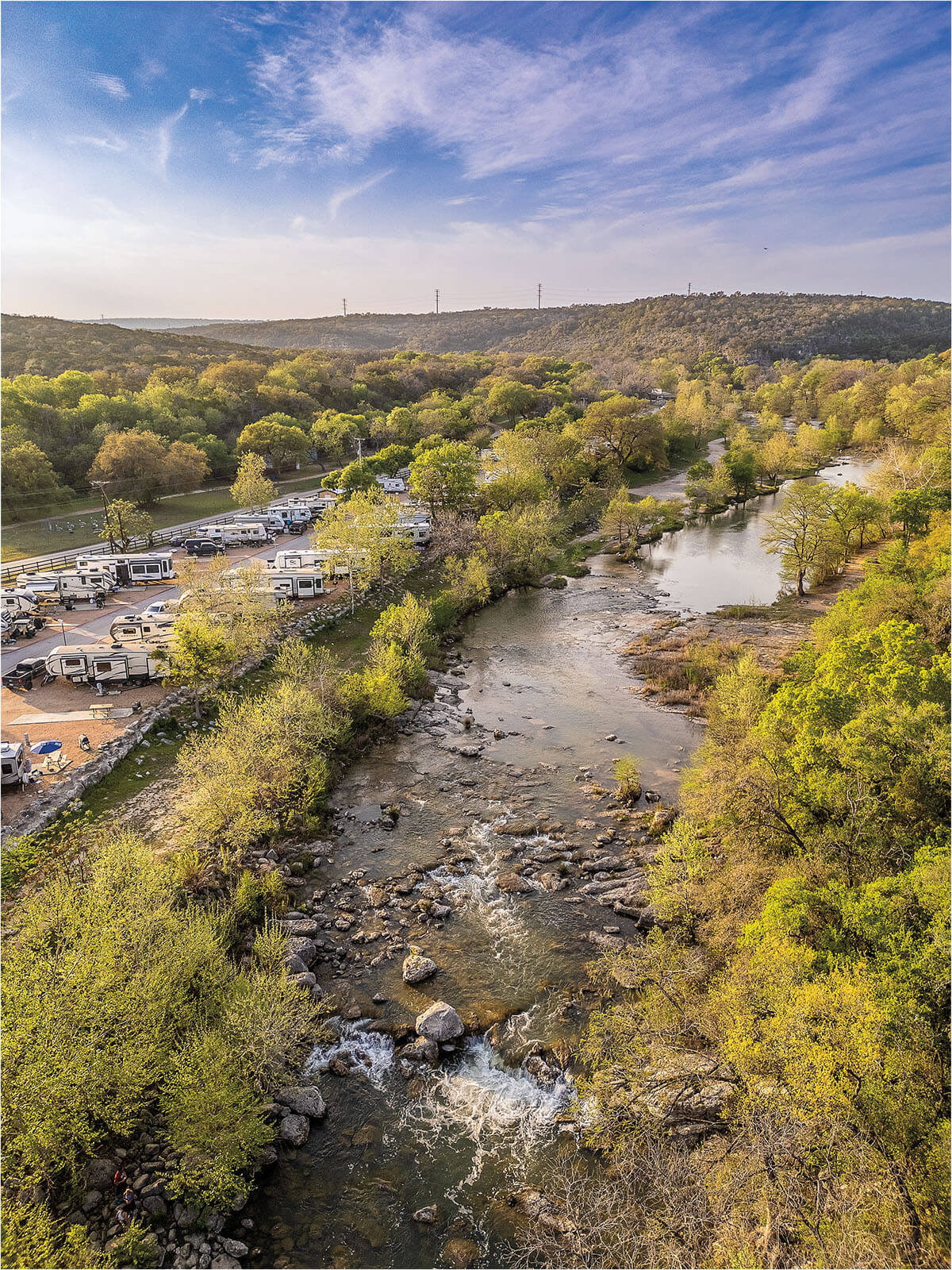 An aerial view of the Guadalupe River under blue sky, with trees flanking its bank. In the left of the image, numerous white RVs are parked in a small parking lot