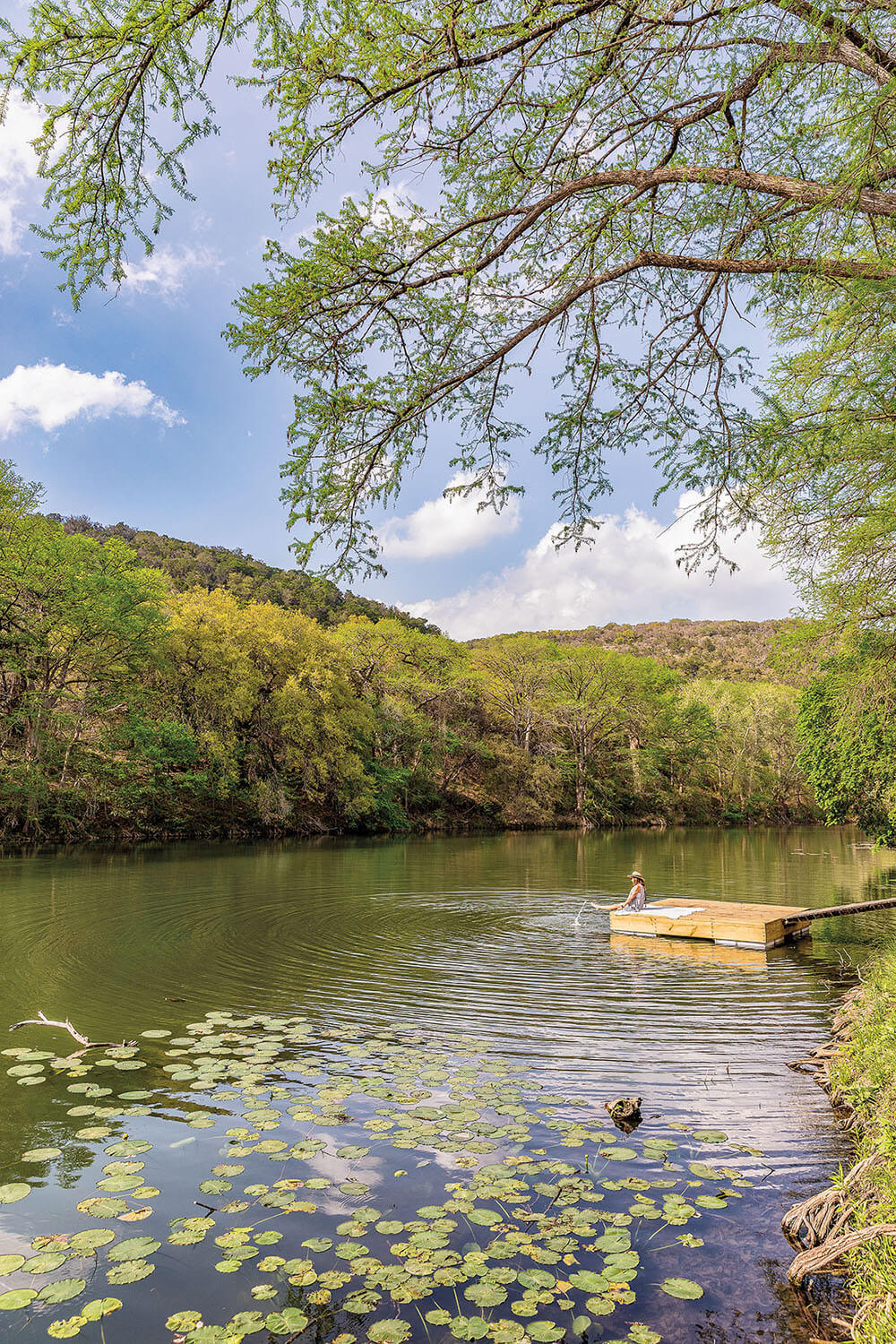 A person sits on a wooden dock with their feet in the water along a greenish-blue river near lots of green trees and lily pads