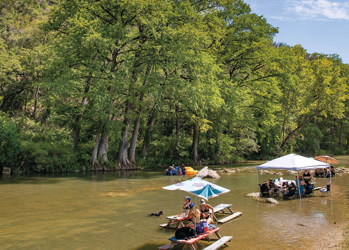 People sit at picnic tables that have been placed in the Guadalupe River, underneath wide umbrellas