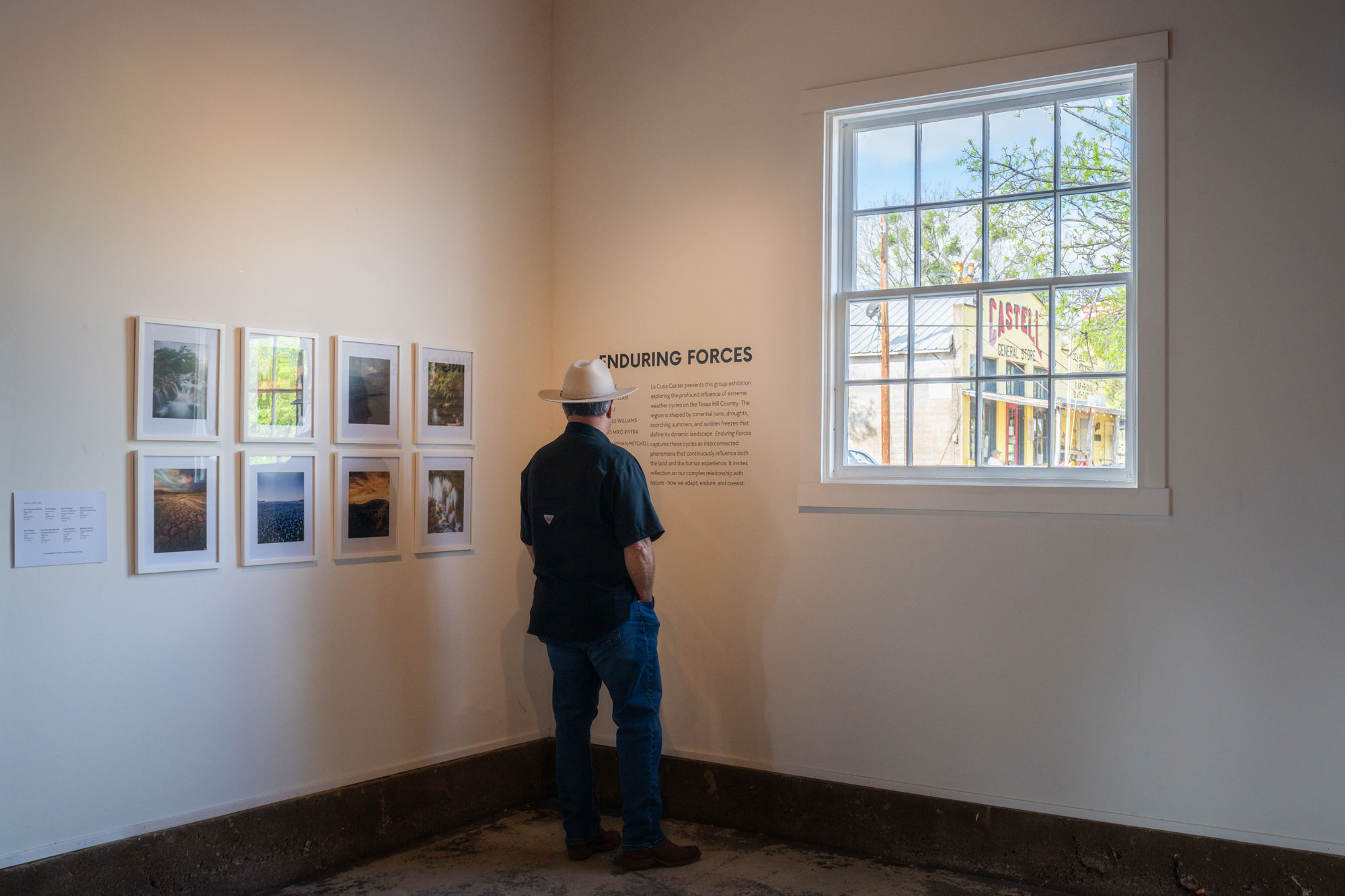 A man in a hat stands in a white room looking at framed photographs on a wall and a text display that reads, "Enduring Forces."