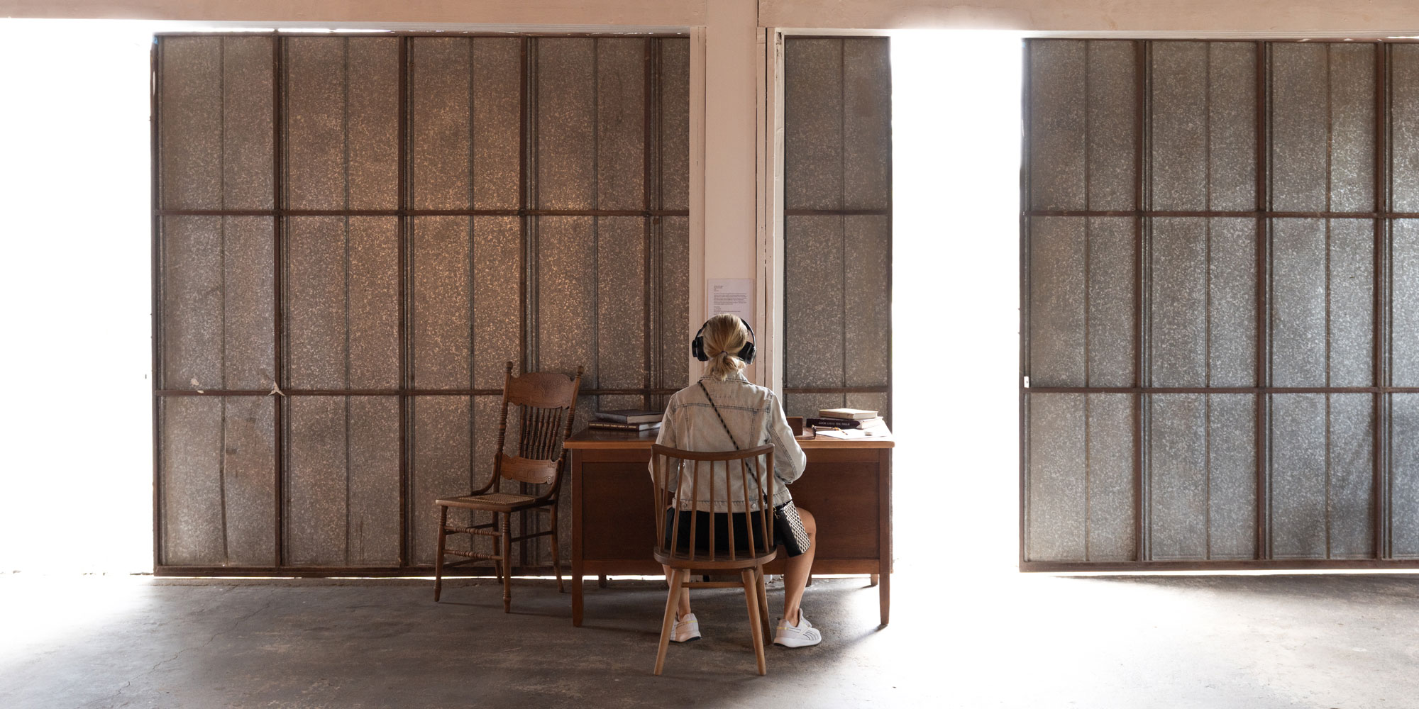 A woman faces away from the camera and sits on a wooden chair while wearing headphones. She faces a wooden desk in a warehouse.
