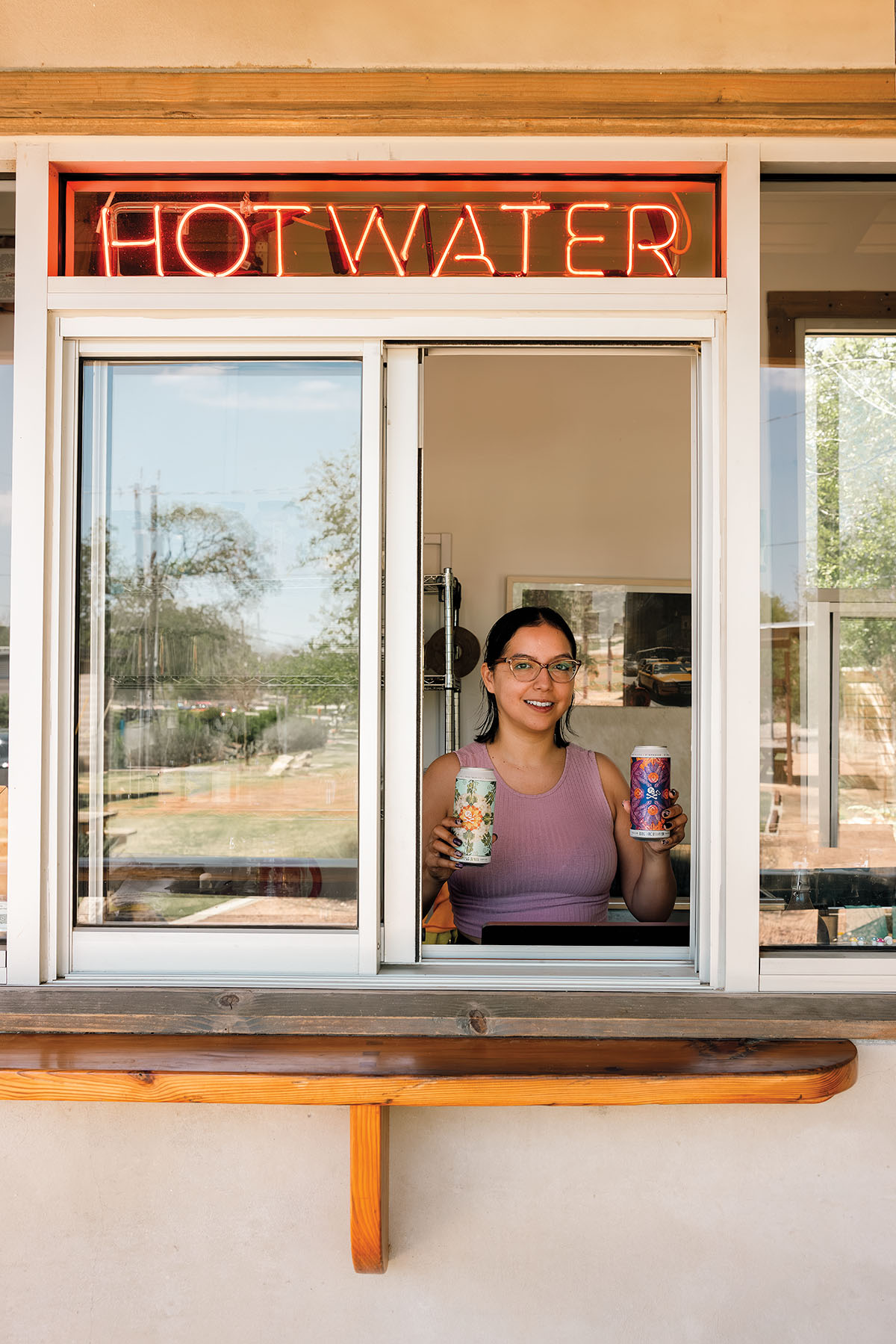 A person holds up two cans at a walk-up service window under neon reading "HOT WATER"