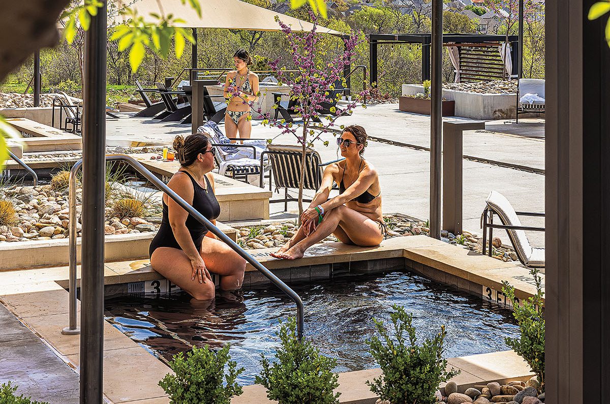 Two people in swimsuits sit partially in a shallow pool under a yellow umbrella. A third person stands behind near a chair.