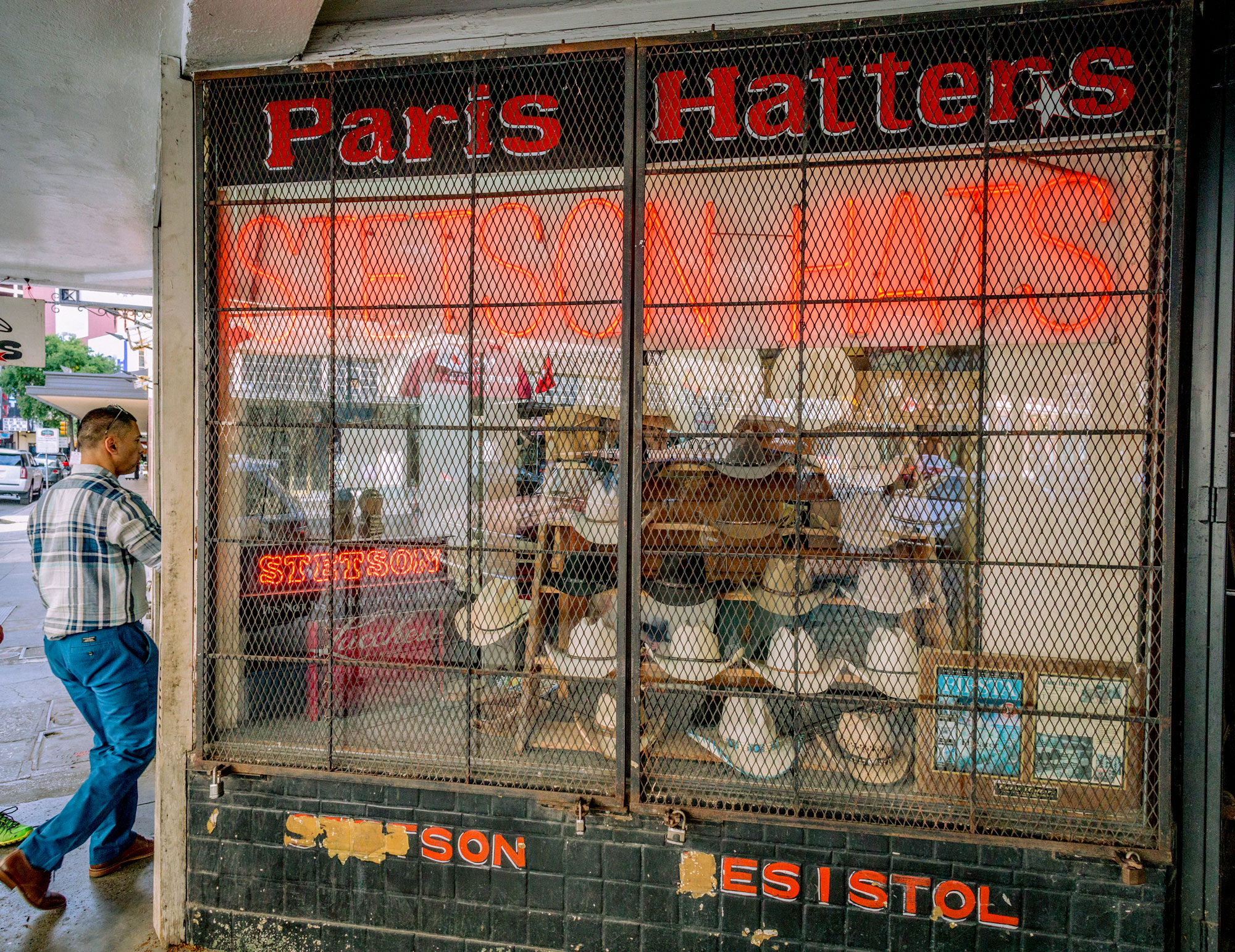 A man walks into a store with a front window filled with cowboy hats. The sign reads "Paris Hatters" with a neon "Stetson" underneath.
