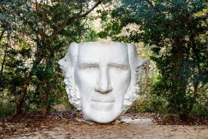 A large white statue head of a man with long hair sits on the dirt ground surrounded by green forest