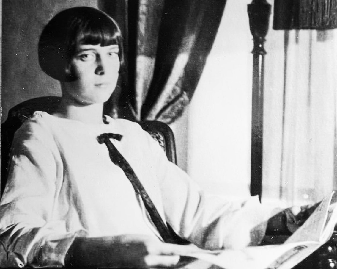 A black-and-white image of a woman sitting in a chair, holding a pamphlet.