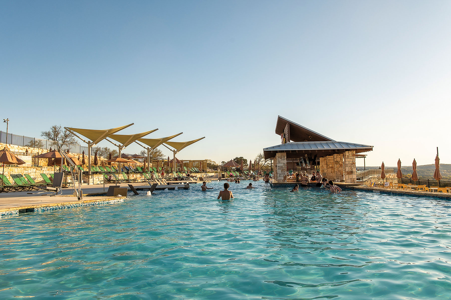 A group of people swim and wade in bright blue pool water under sunny skies