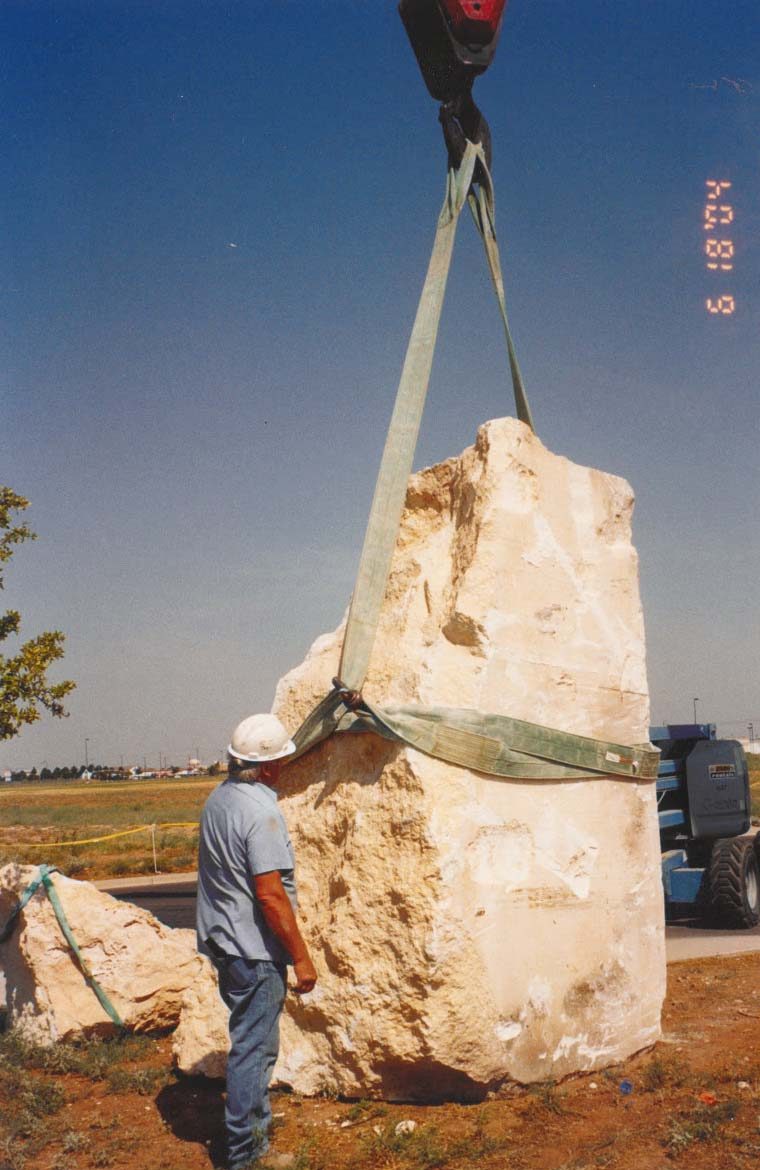 A person in a blue shirt, jeans and a white hard hat stands next to a large piece of white limestone being lifted by a gray construction crane