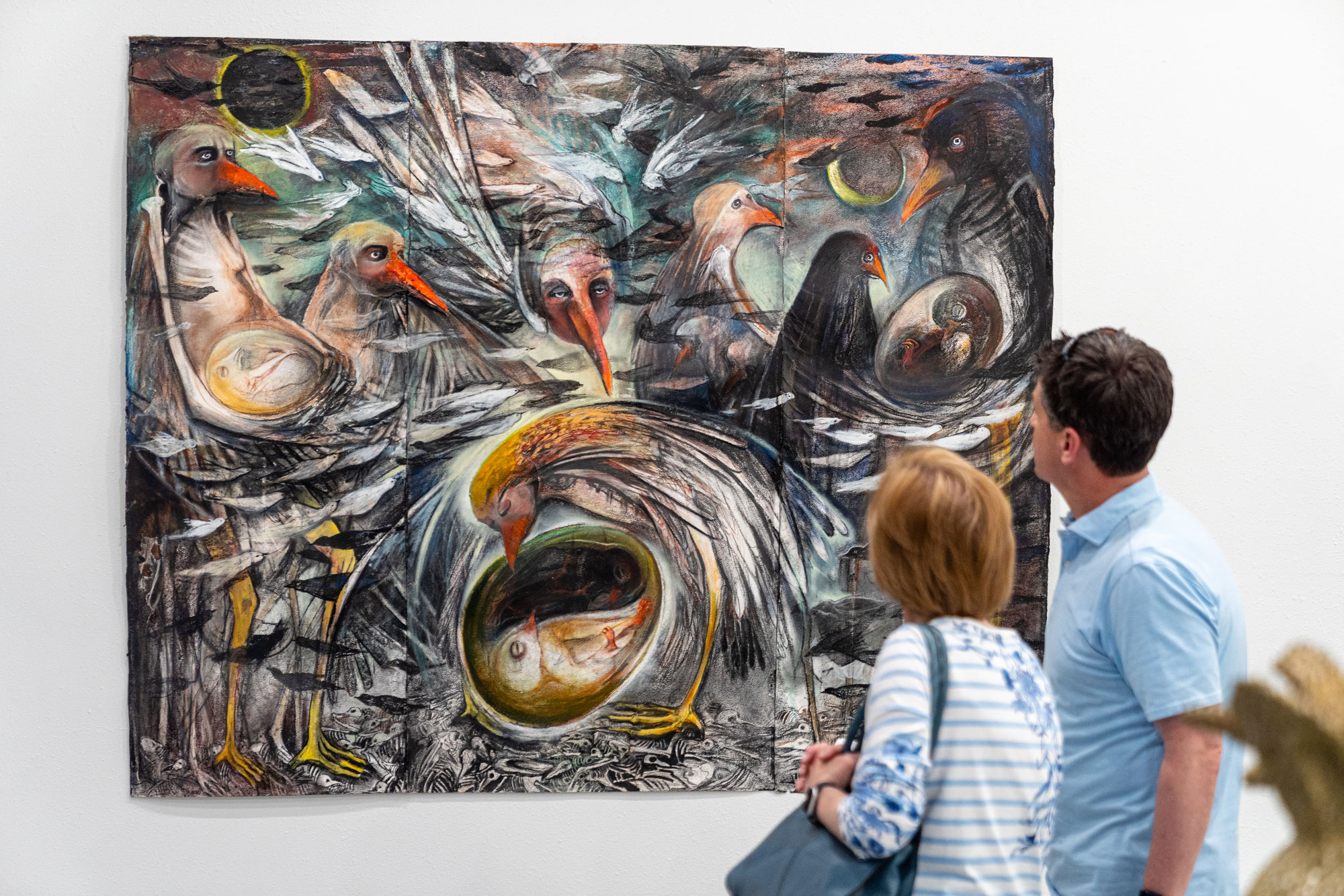 A man and woman look at a painting on the wall. The painting depicts several bird-like figures with humanoid eyes and sullen expressions in muted gray, blue, and orange colors.