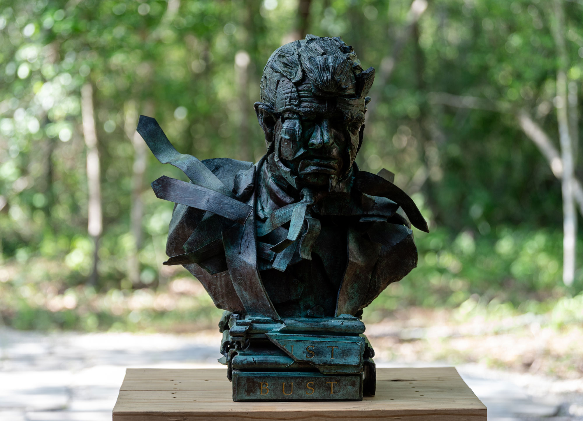 A bronze bust of a grimacing man sits on a platform outside with trees in the background