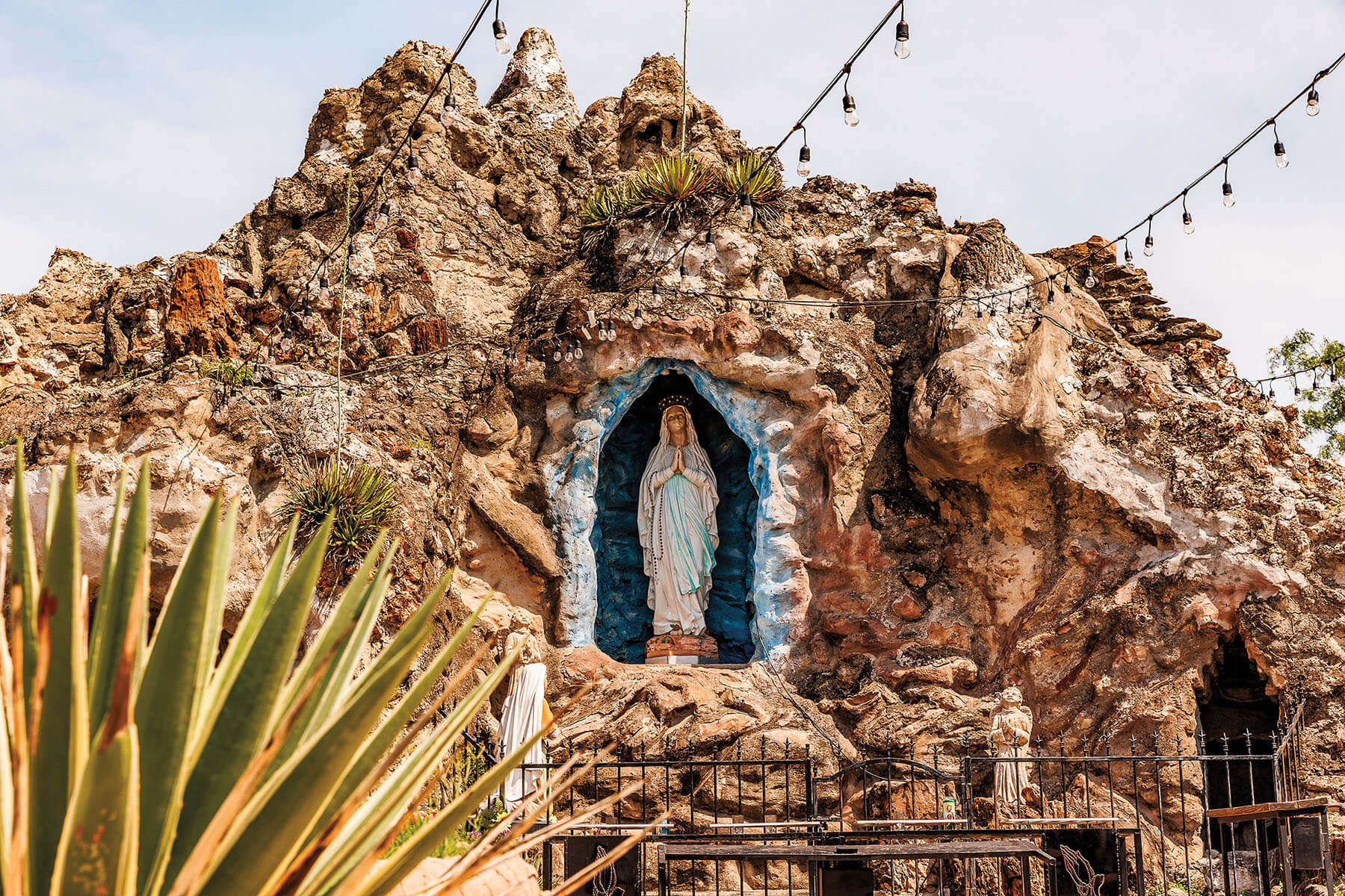 A blue-painted religious symbol built into a rock outcropping. Black-corded string lights adorn the figure, which is behind a fence and next to pointy green cacti