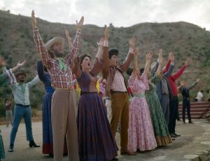 Actors dressed in colorful pink, orange, green and blue Western wear costumes pose on the beige stage surrounded by greenery and brown cliffs.