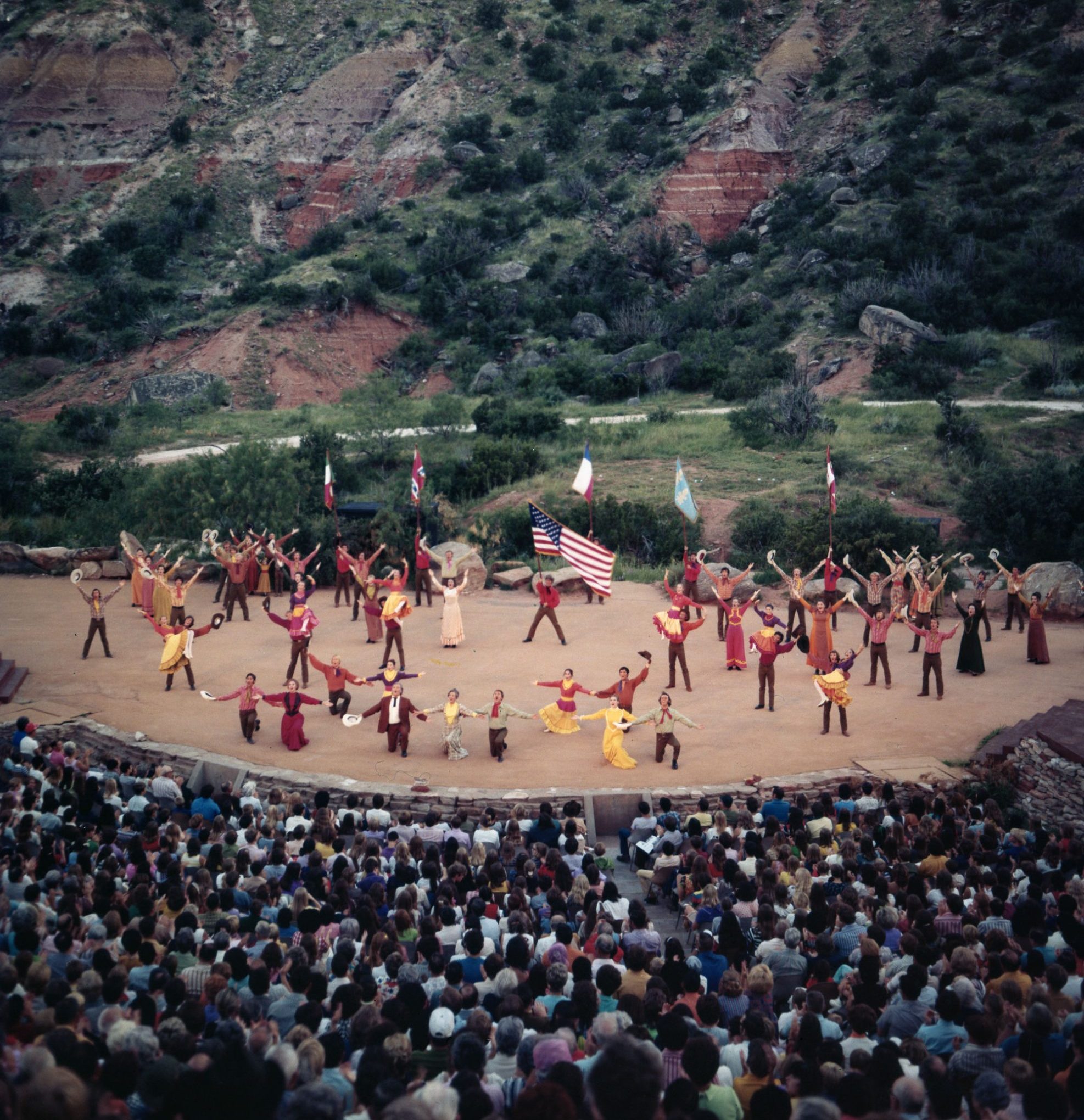 A distant, birds-eye view of an amphitheater filled with actors and audience members surrounded by cliffs