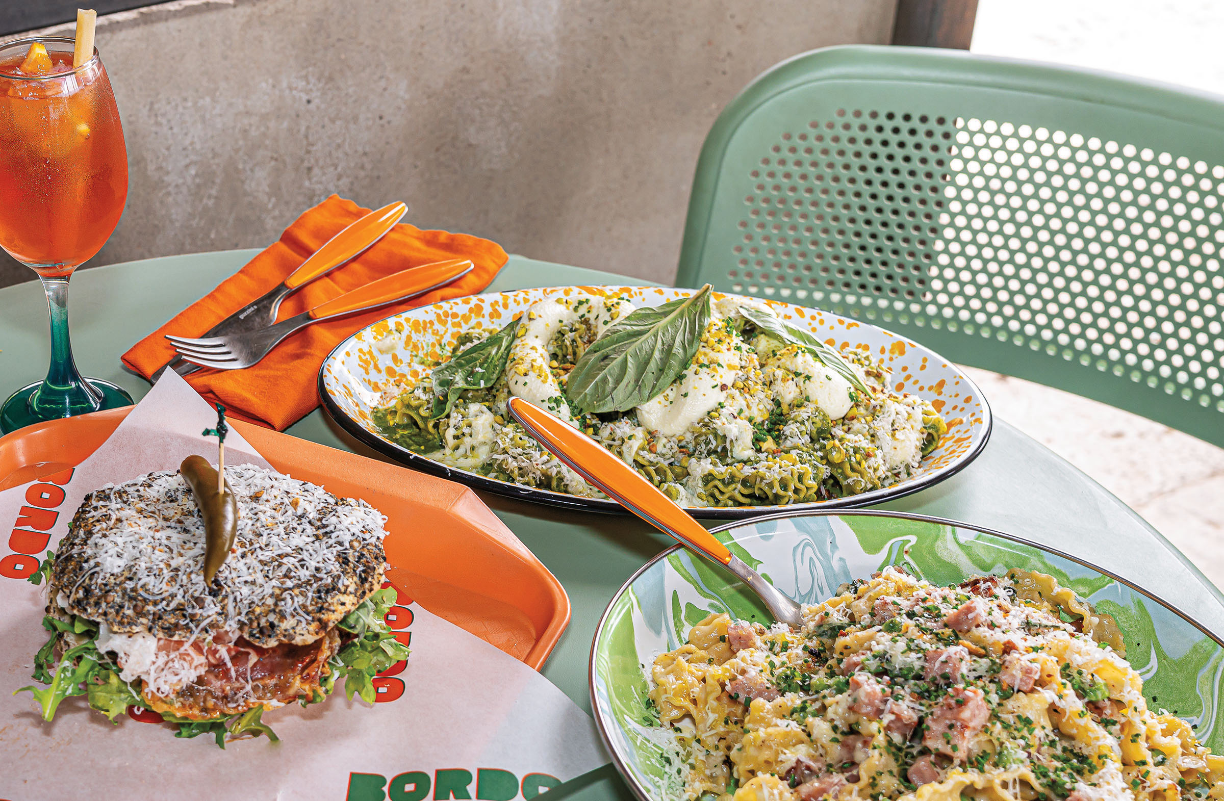 A sandwich and two pasta dishes are set on a table next to an orange, icy drink.