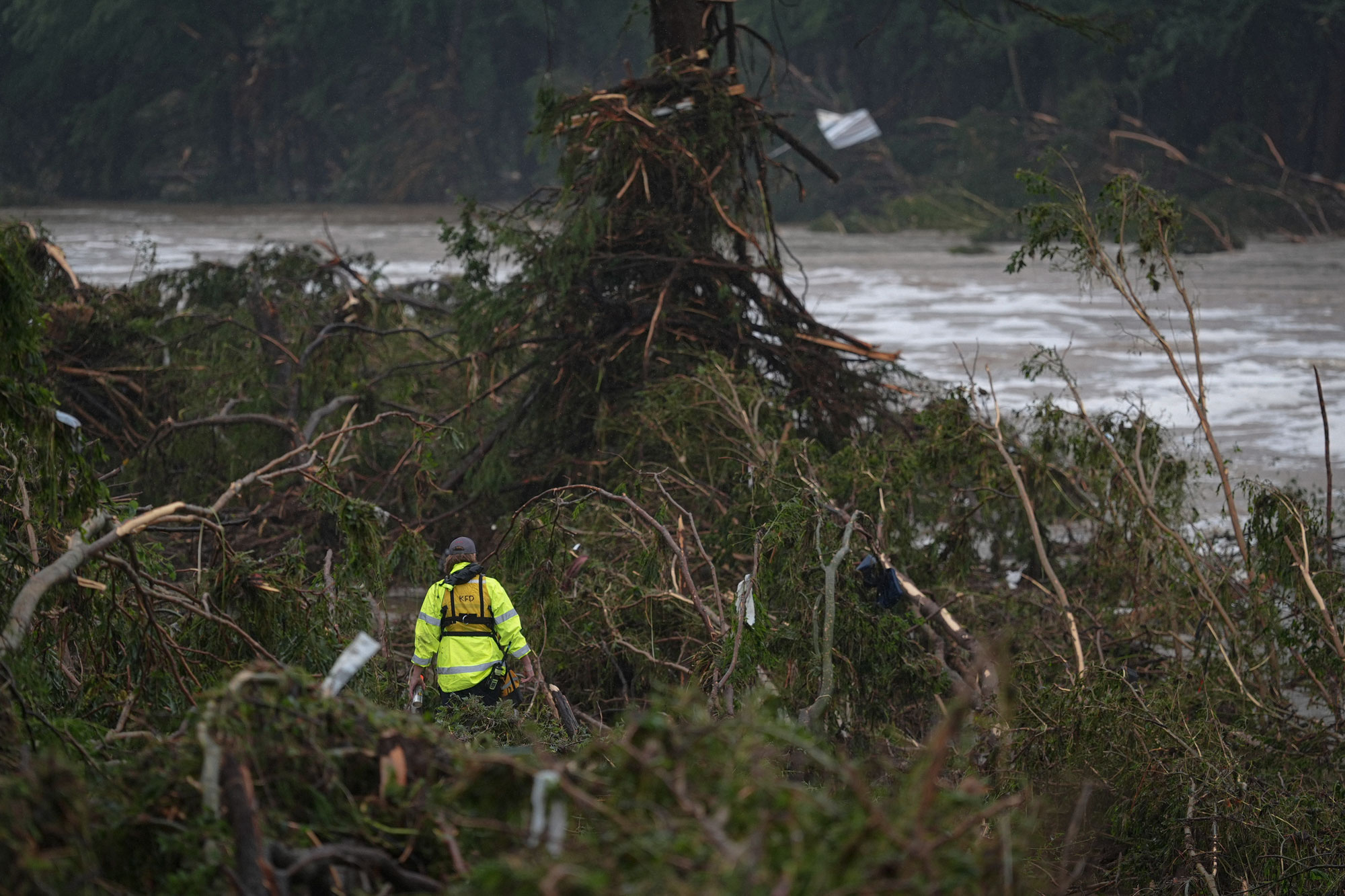 A man in a bright yellow first responder jacket walks through debris in front of a flowing river.