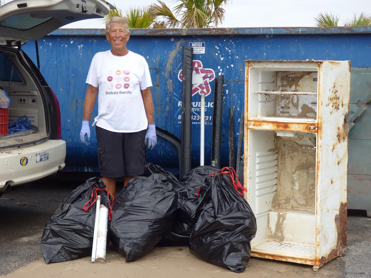 A man in a white shirt, shorts, and wearing gloves stands next to an open SUV trunk next to several full trash bags.