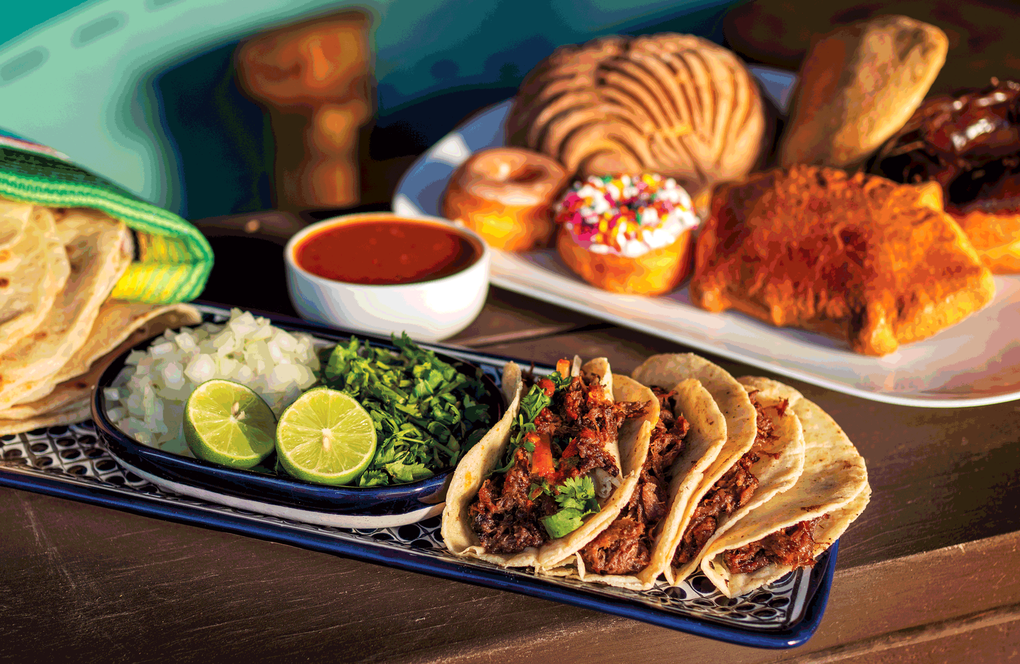 A plate of four soft tortillas stuffed with beef, next to a plate of chopped onions, sliced limes, and cilantro