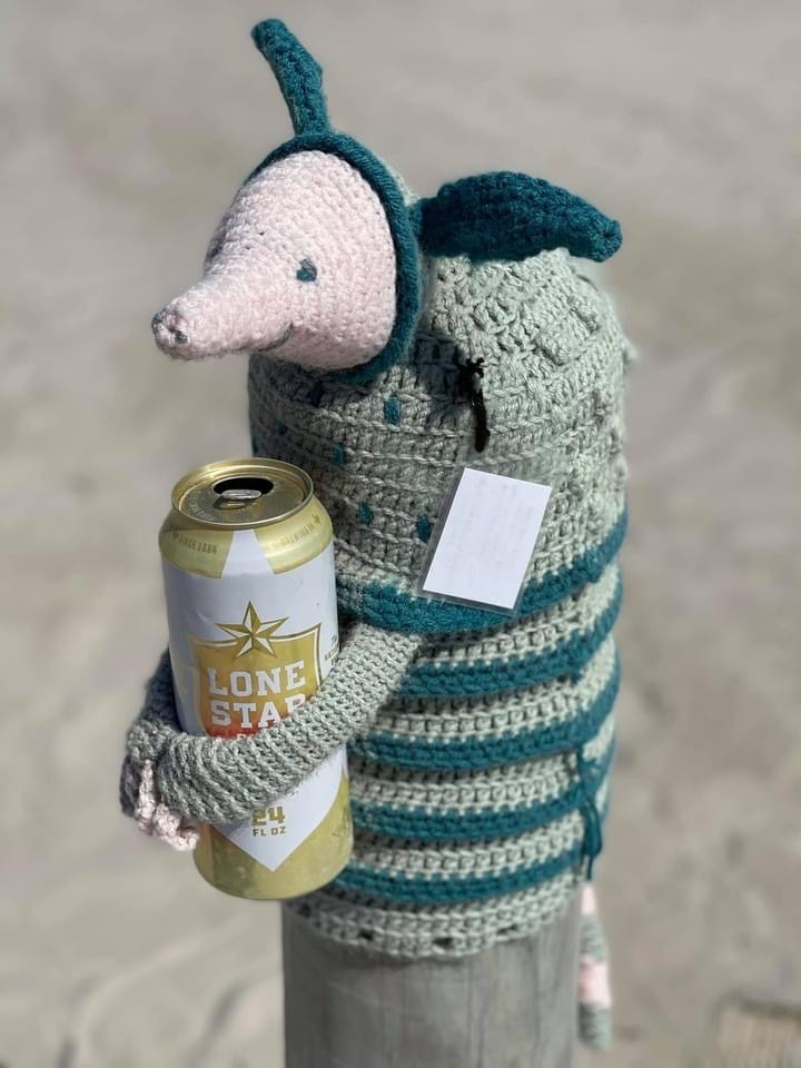 A crocheted Arnie the Armadillo holding an empty Lone Star beer can on the shore of Port Aransas' Nueces County beach.