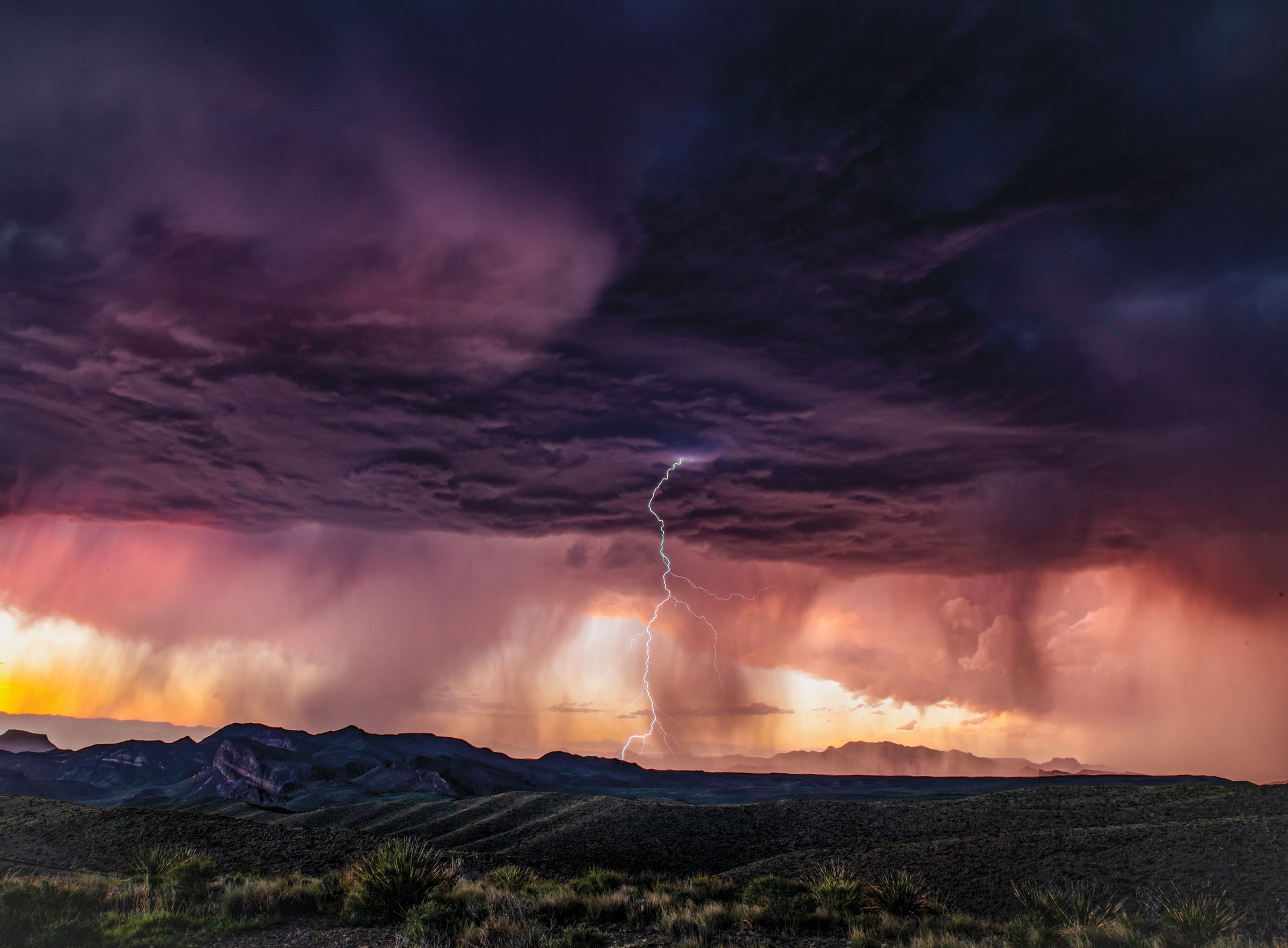 A stormy skyscape with a purple and black cloud overhead, rays of orange light coming down, and a silhouette of a mountain 