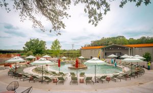 Several green patio lounge chairs and red-orange umbrellas surround a circular pool with a fountain in the center. Buildings surrounded by foliage are in the background.