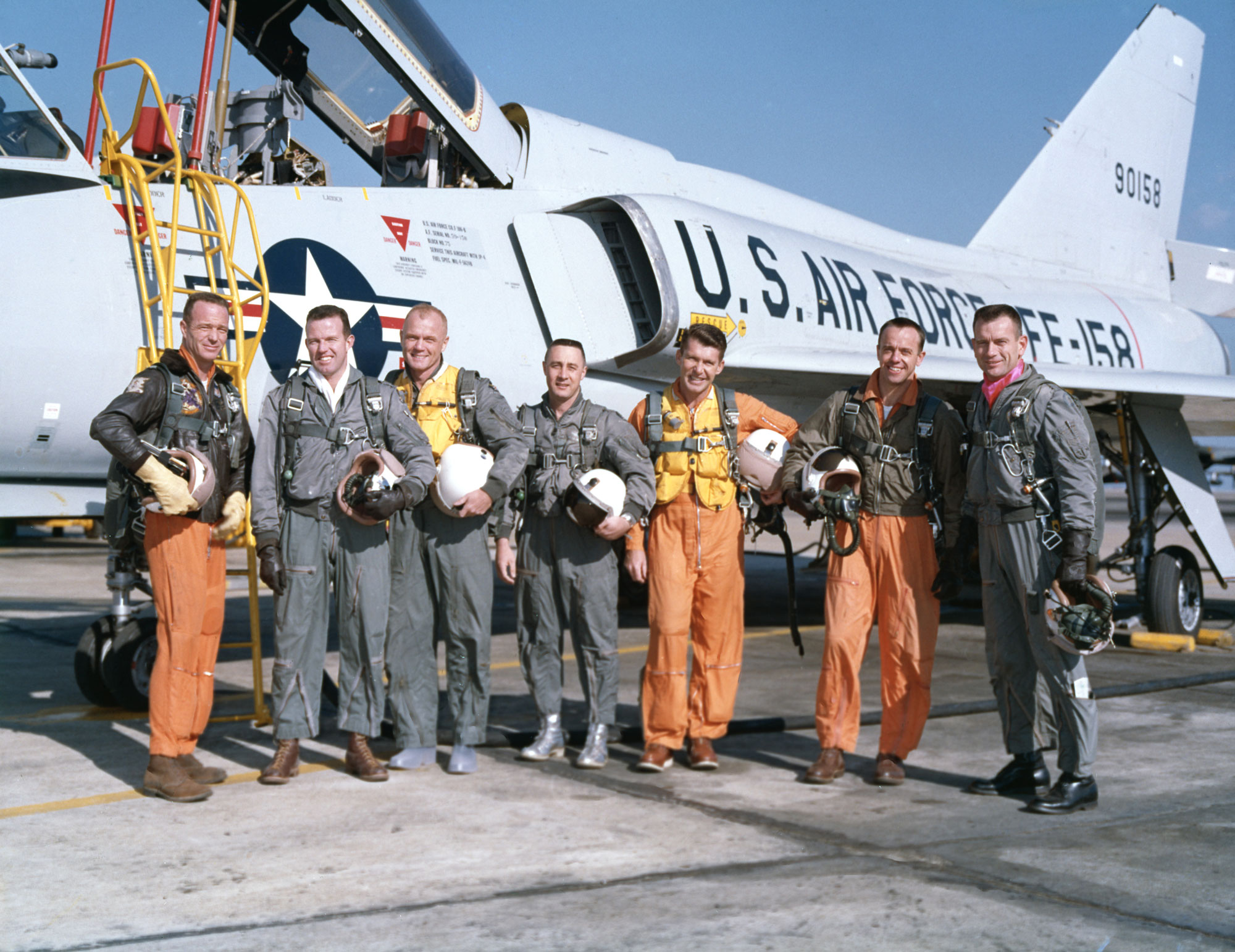 Seven men dressed in green and orange air force uniforms stand in front of a white U.S. airforce plane.