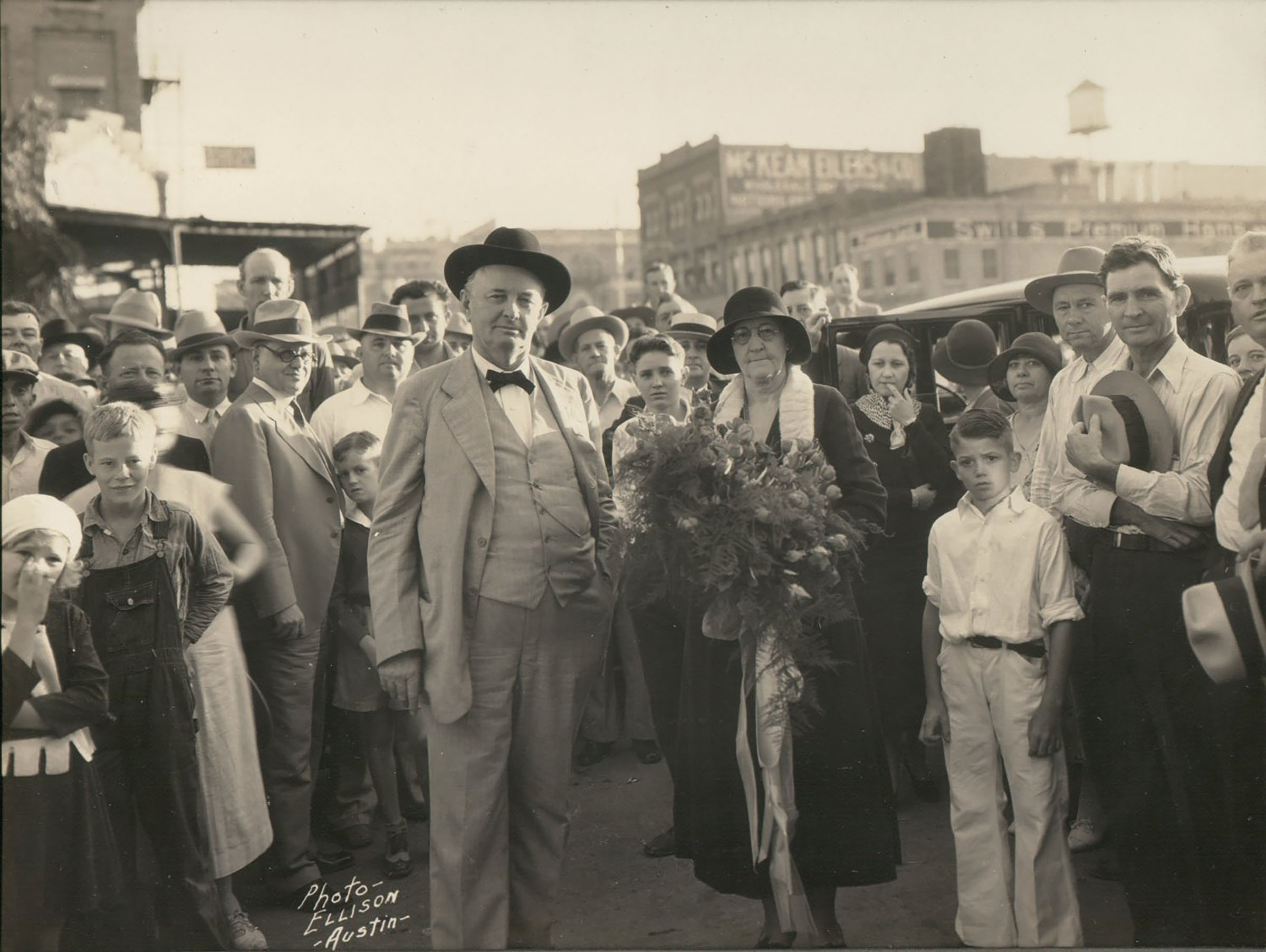 An old black and white photo of a an older man and woman holding a bouquet of flowers standing in front of a crowd of people on a town street.