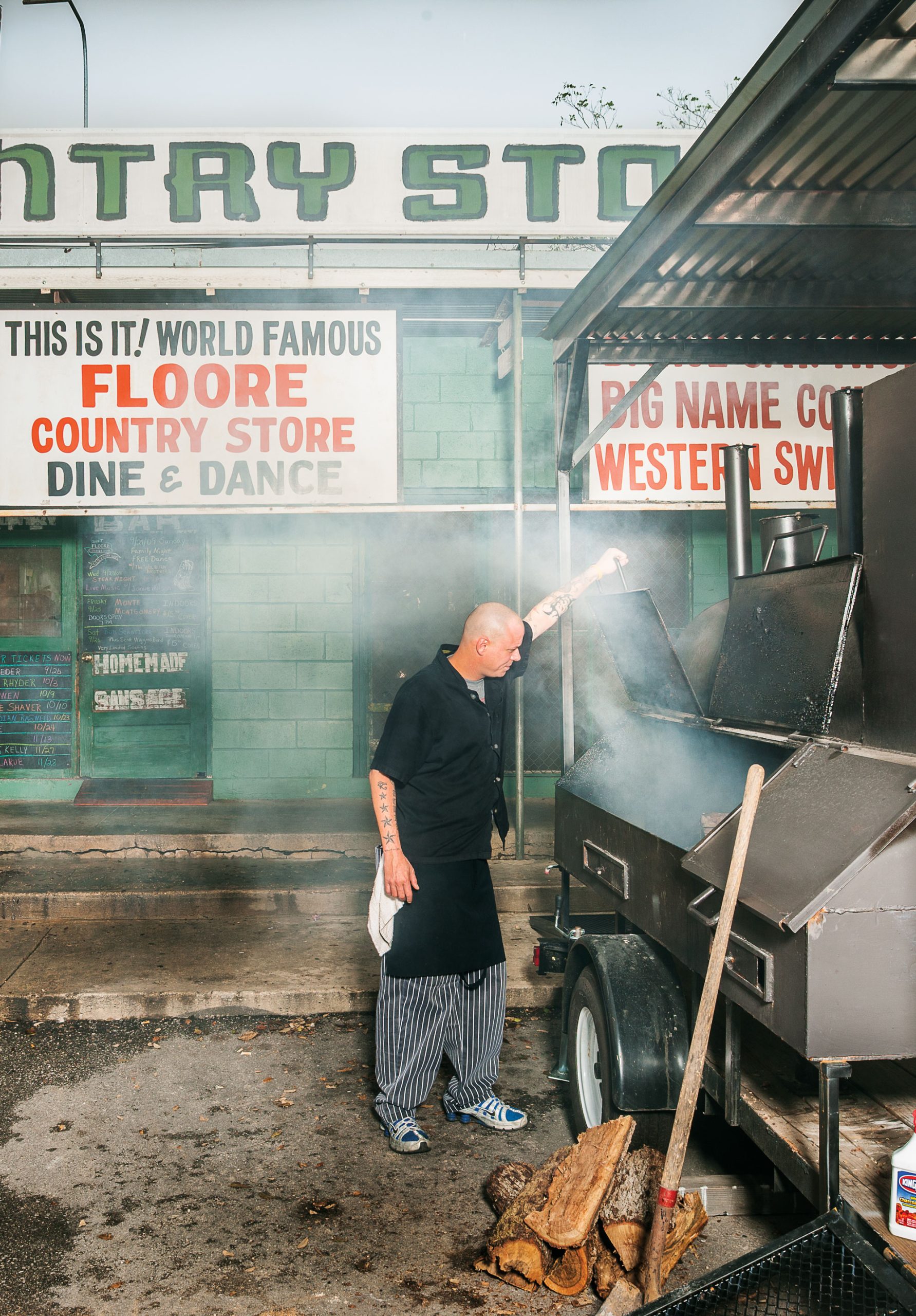 A man in a black shirt and jeans checks on a smoking barbecue pit in front of a green building and hand-painted signs
