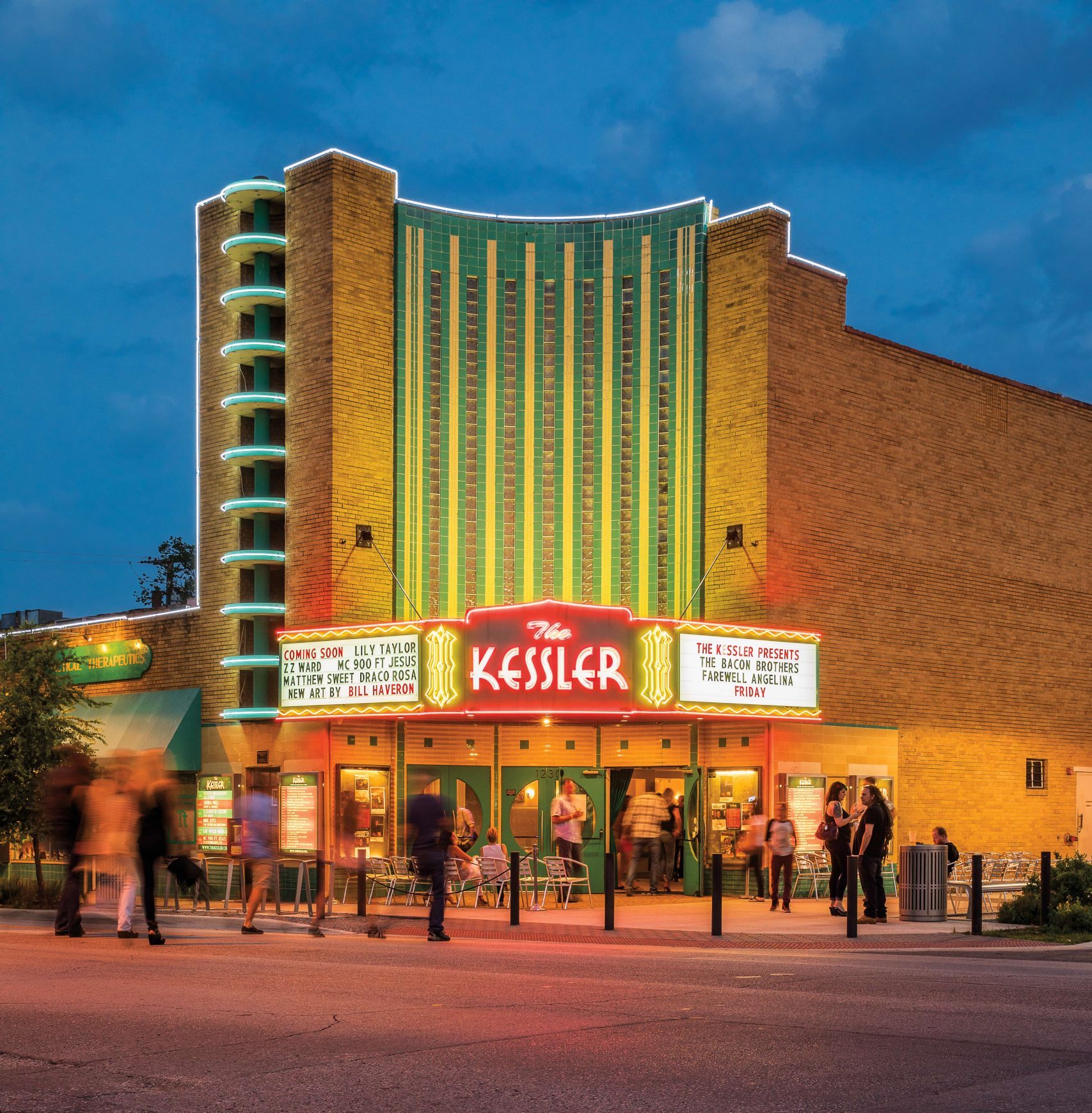 People walk into a lit up theater at dusk that has green neon lights going up and down the front of its facade and the sign reads "The Kessler"