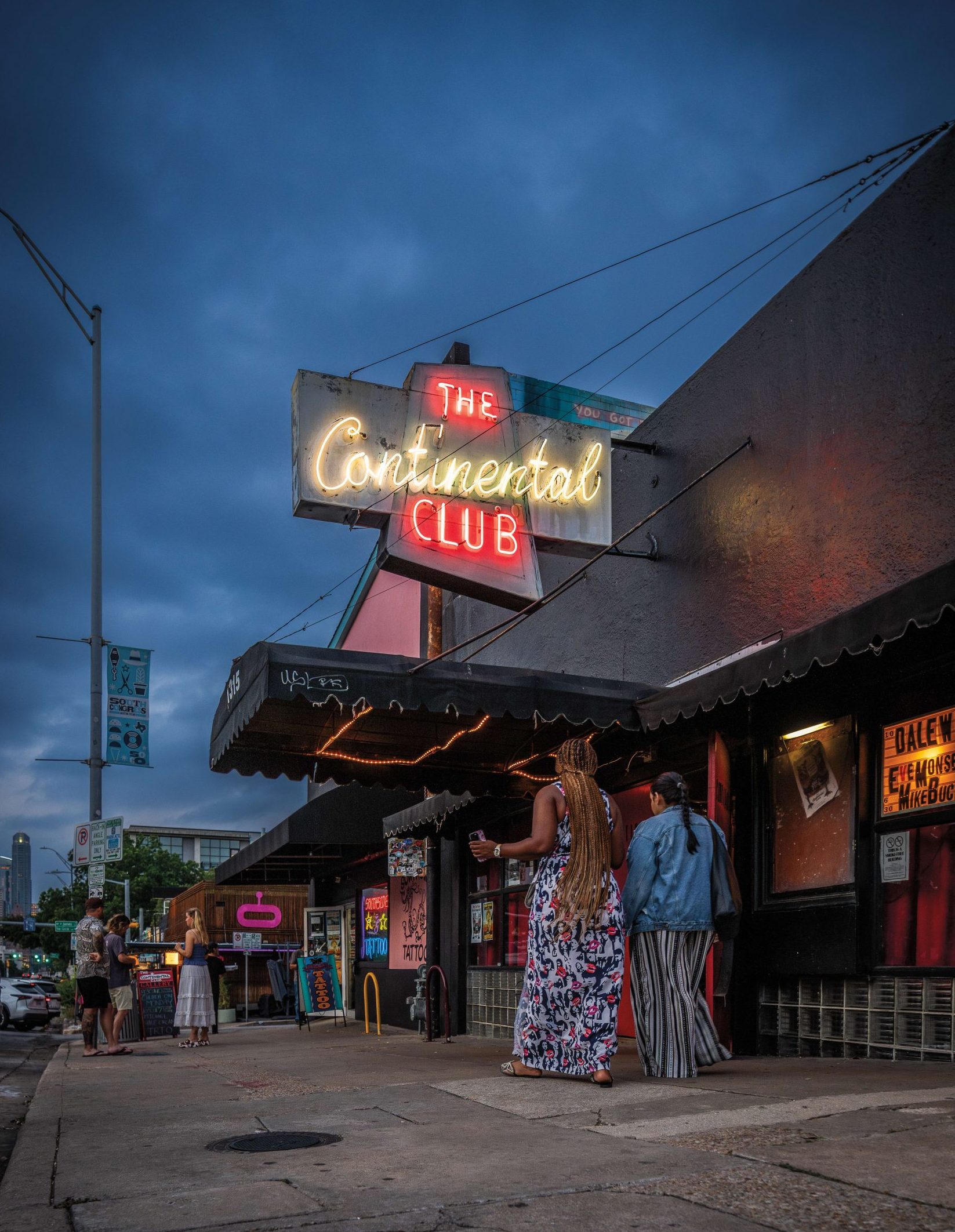 People walk by a building facade under a neon light that reads "The Continental Club"