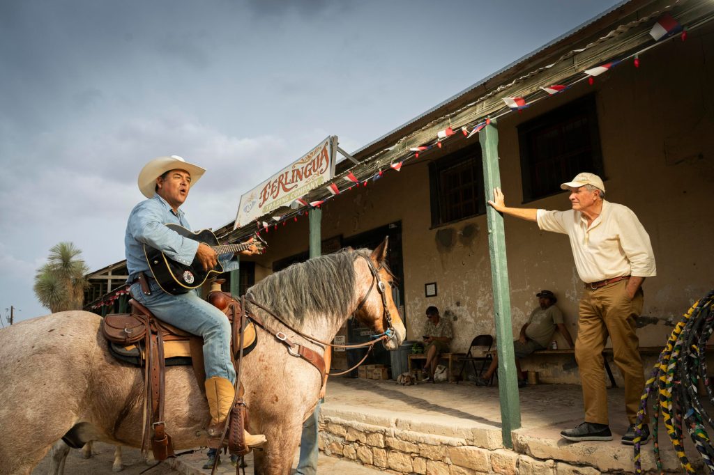 A man in a cowboy hat and riding a horse holds a guitar and sings in front of a rustic porch and another older man.