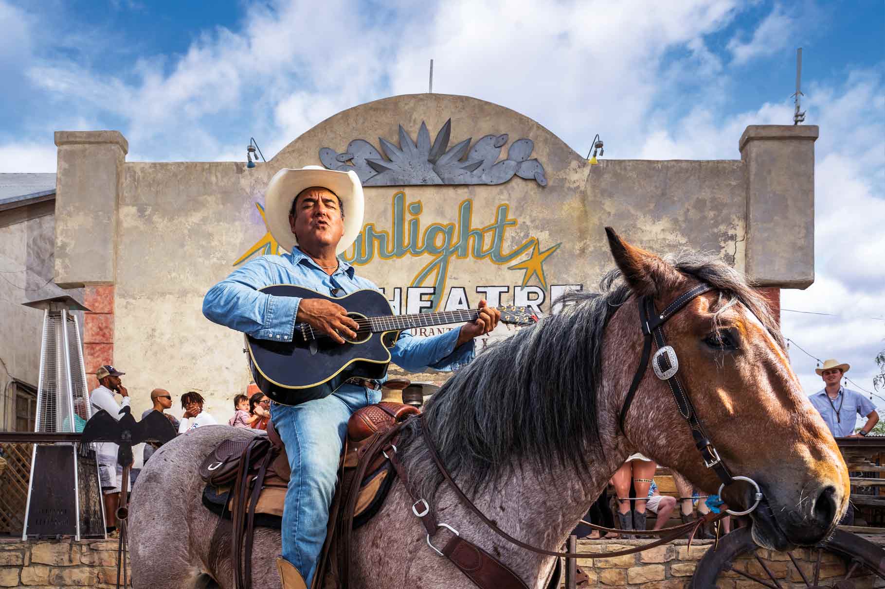 A man dressed in denim and a cowboy hat sits on a horse while holding a guitar and singing. They are in front of an old building that has a hand painted sign that reads "Starlight Theatre"