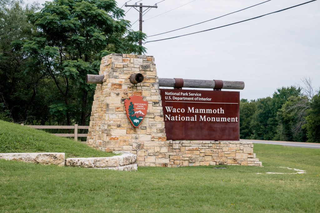 A brow sign that reads "Waco Mammoth National Monument" in white letters next to a National Parks symbol stands on a green lawn.