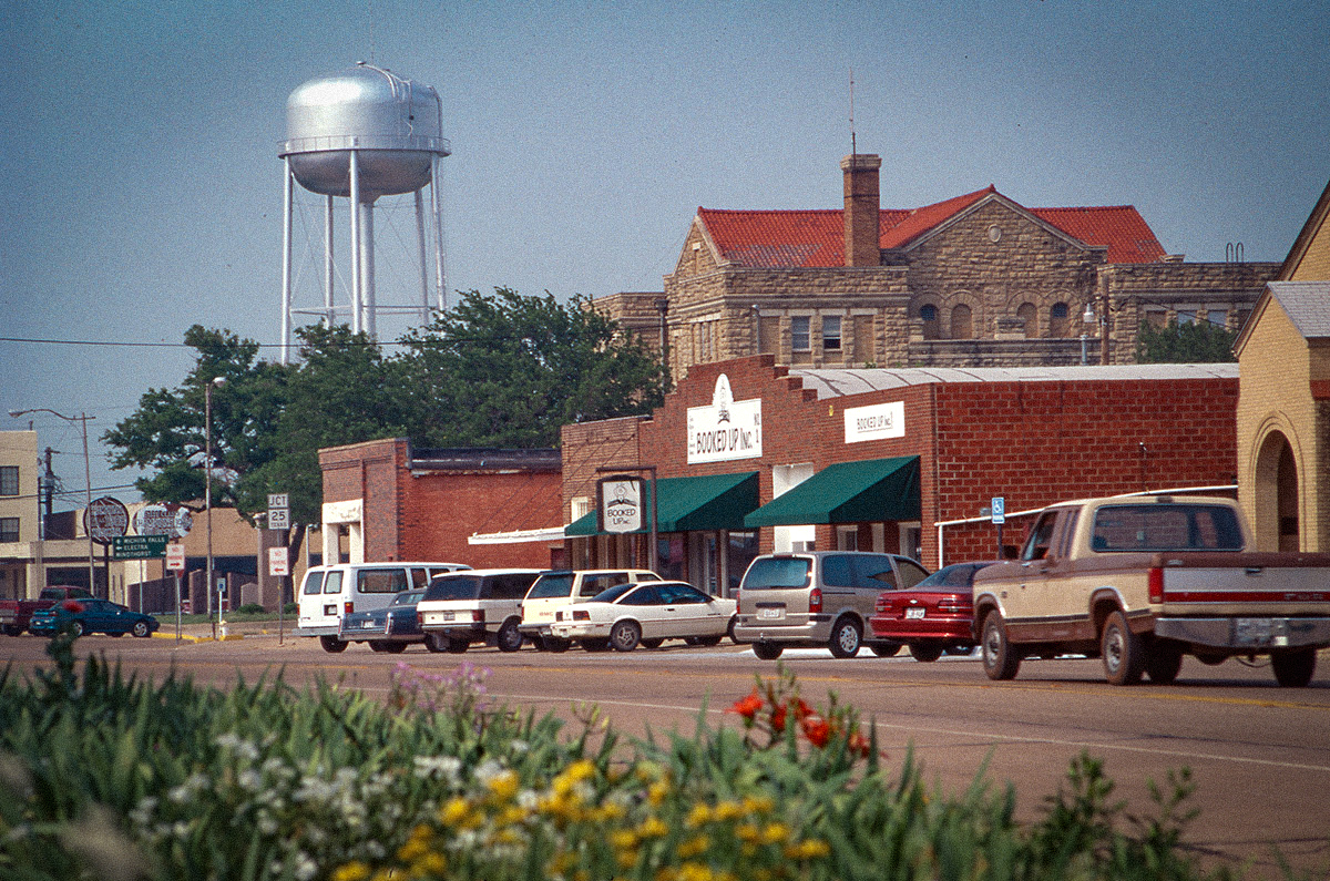 Cars are parked in front of brick stores along a grey road