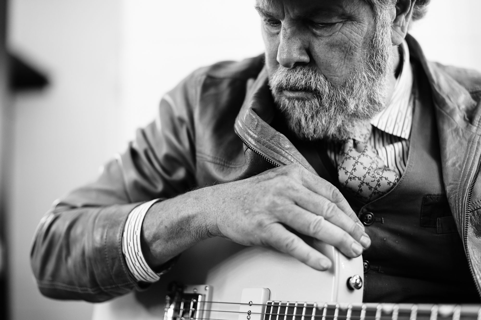 A black and white photo of an older man with a beard dresed in a leather jacket sitting and somberly looking down with a guitar in his lap