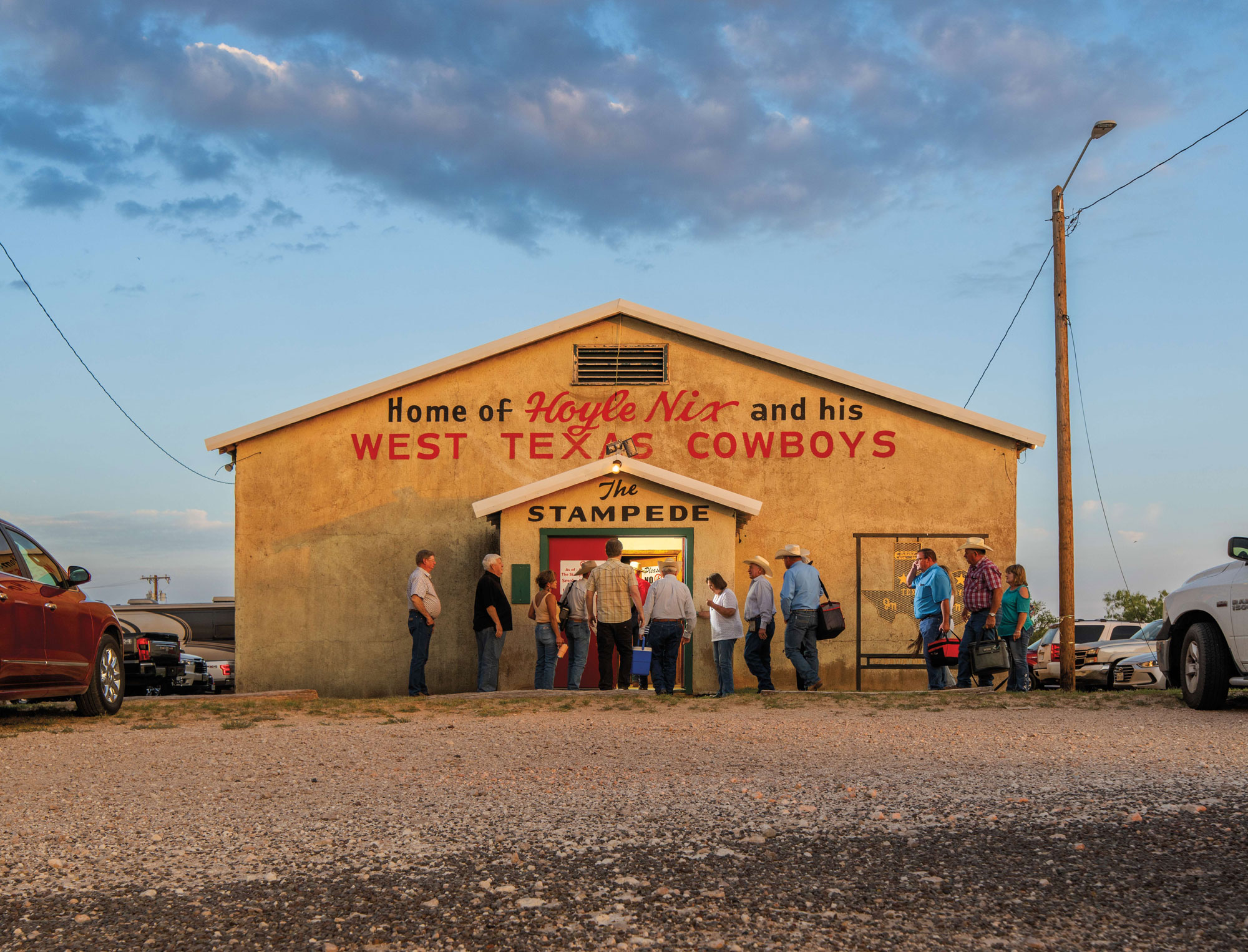 A crowd of people dressed in Western dance wear enter a simple beige building with red and black words painted on its front that read "Home of Hoyle Nix and His West Texas Cowboys—The Stampede."