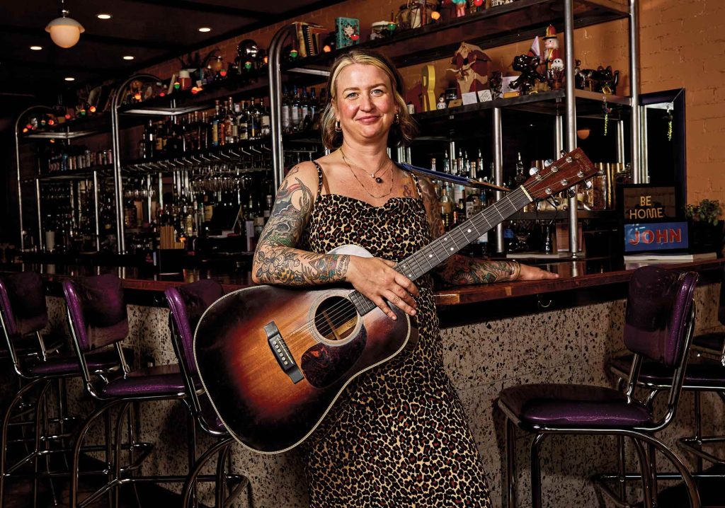 A woman in a dress smiling and holding a sunburst acoustic guitar