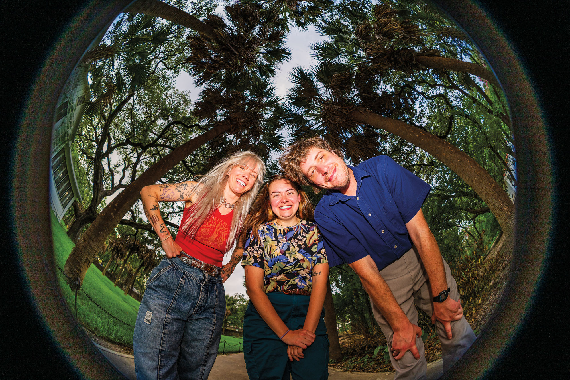 Three people clustered together smile while peering down into a fishbowl camera lens