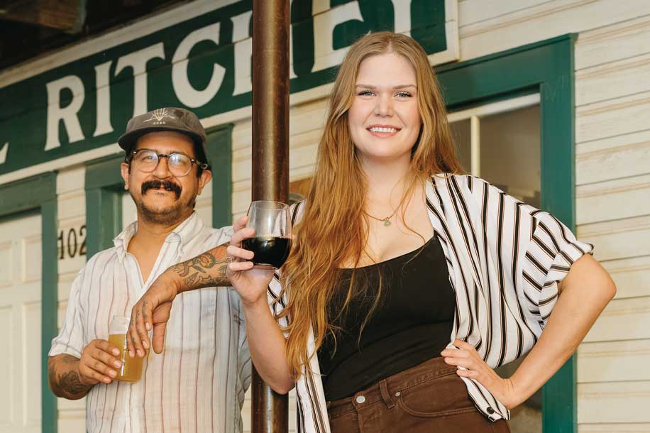 A man and a woman holding a glass of wine pose in front of a storefront with a sign that reads "Ritchey"