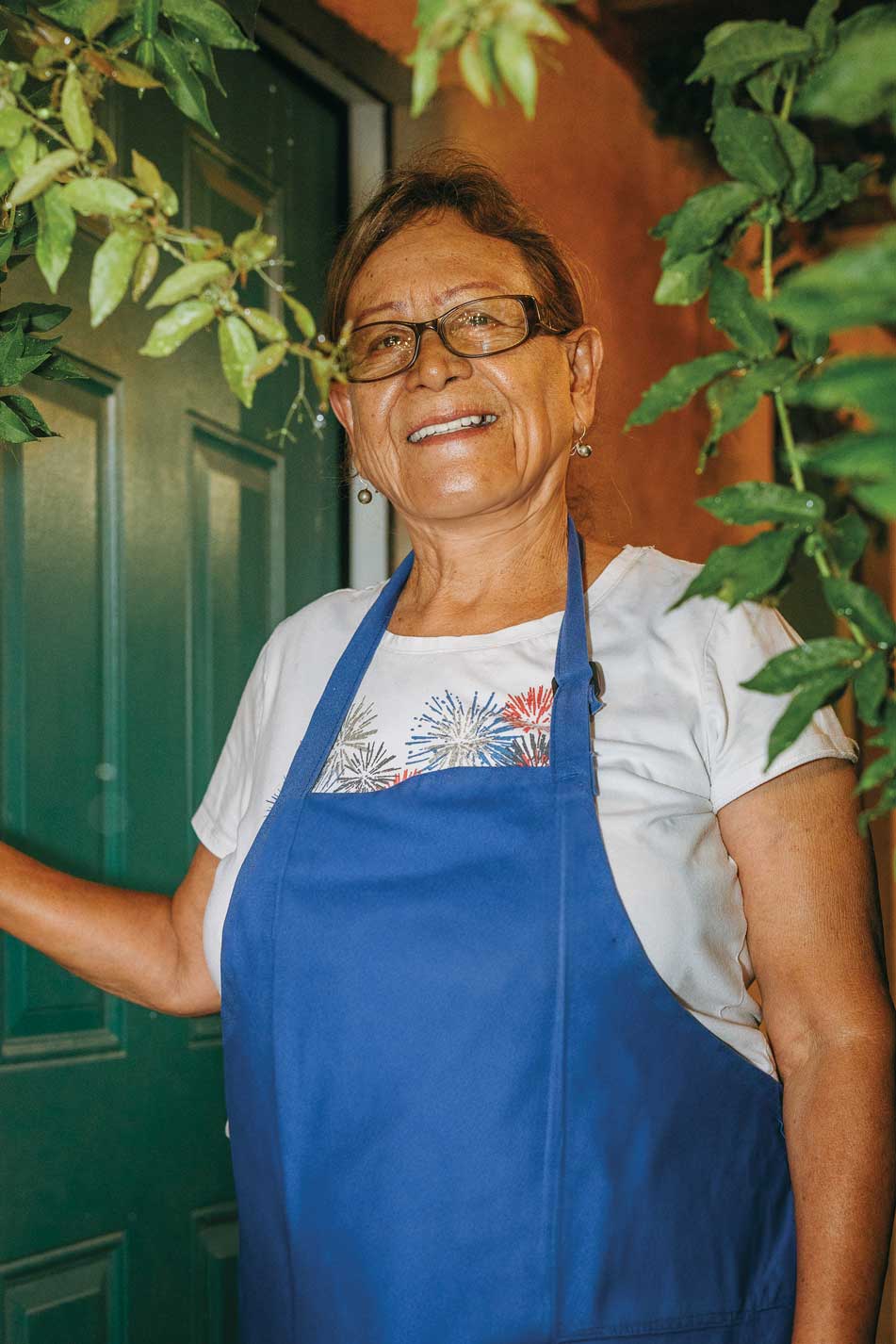 A woman in glasses and a blue apron poses in front of a a green door surrounded by foliage