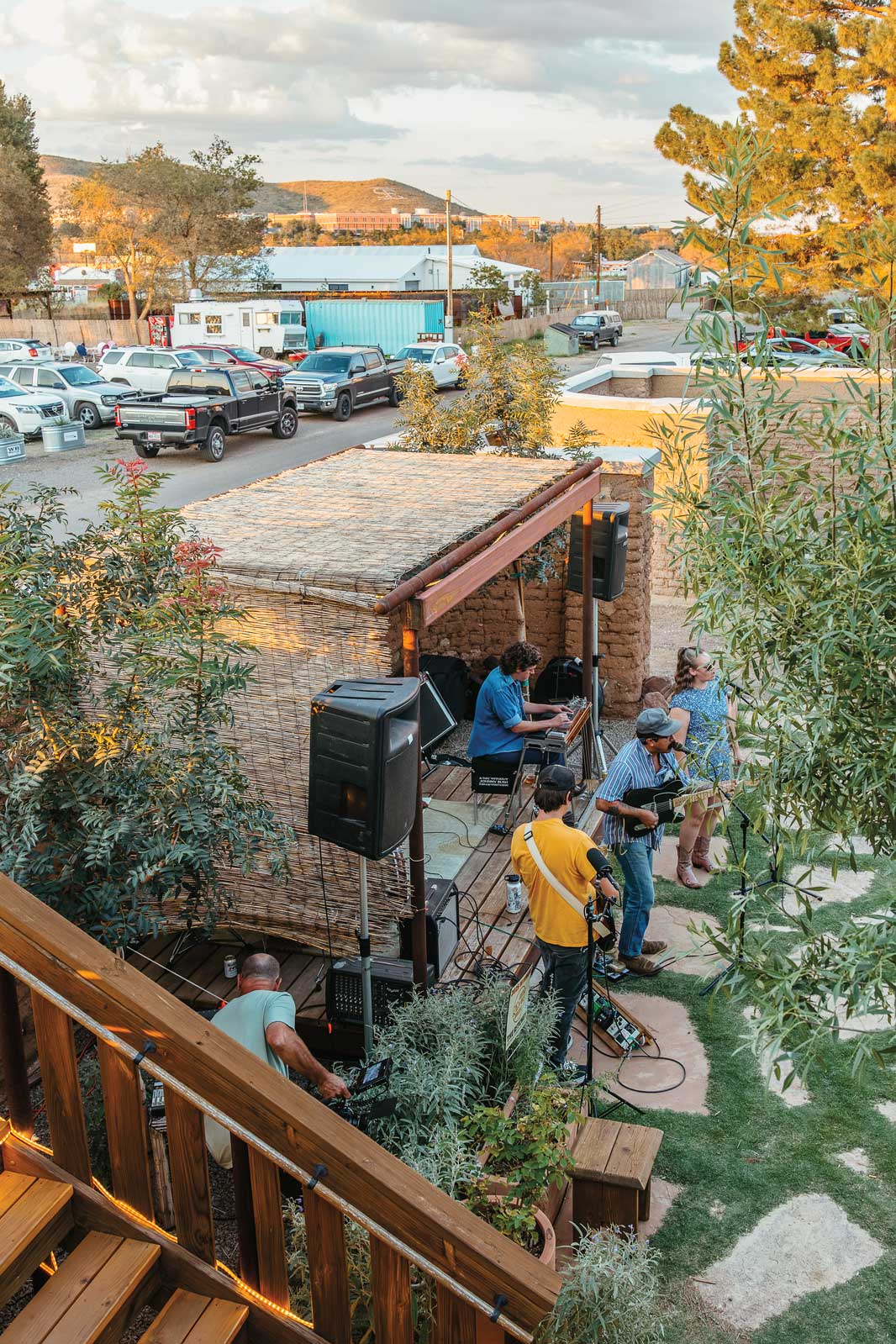 Several people with instruments gather in front of an open stage made of stone in a lawn beside a parking lot