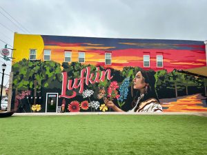 Mural on side of building shows Angelina's face in profile on the right, Lufkin is painted in red on the left, flowers are painted along the bottom and a sunset is painted on top.