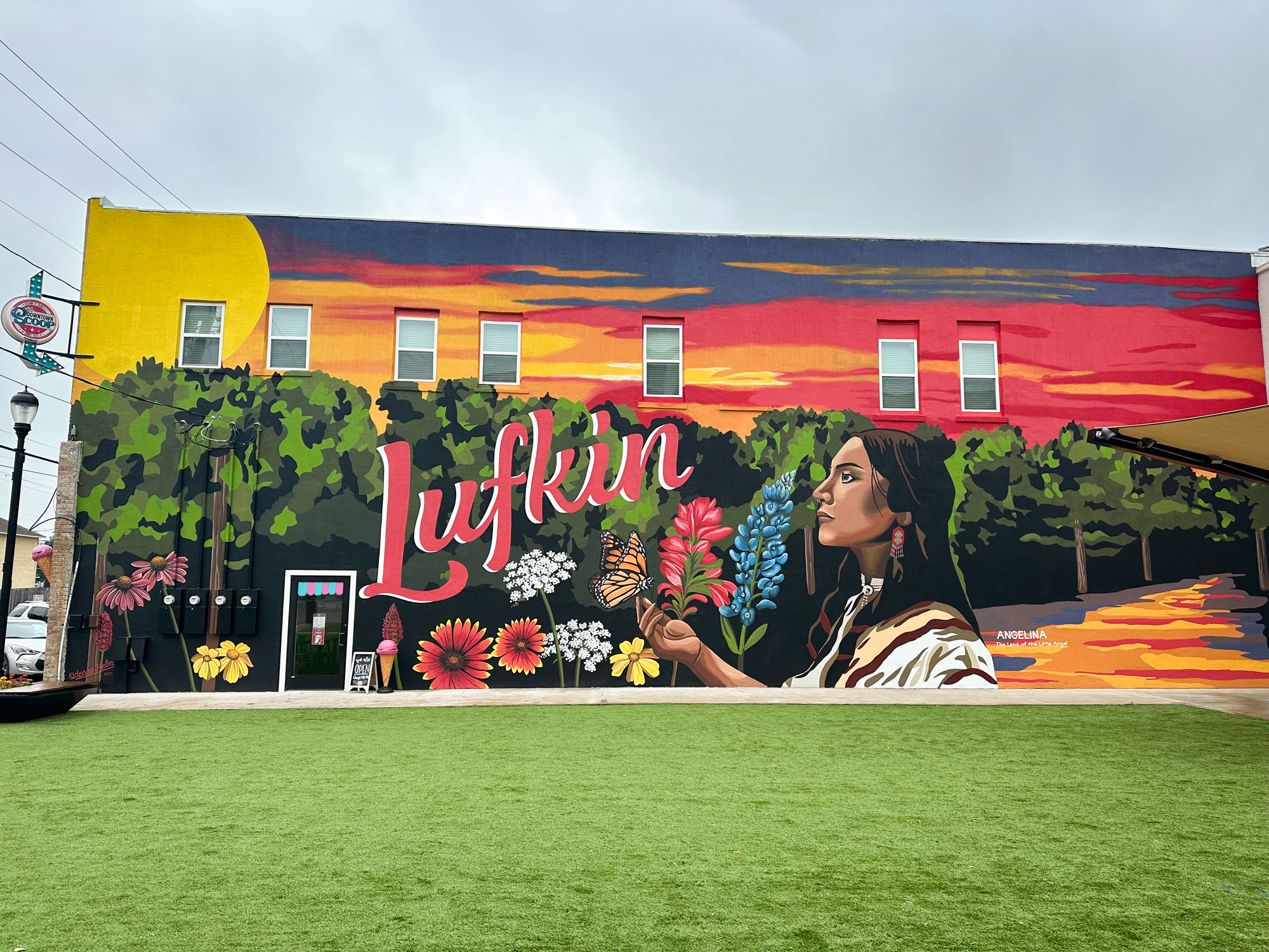 Mural on side of building shows Angelina's face in profile on the right, Lufkin is painted in red on the left, flowers are painted along the bottom and a sunset is painted on top.